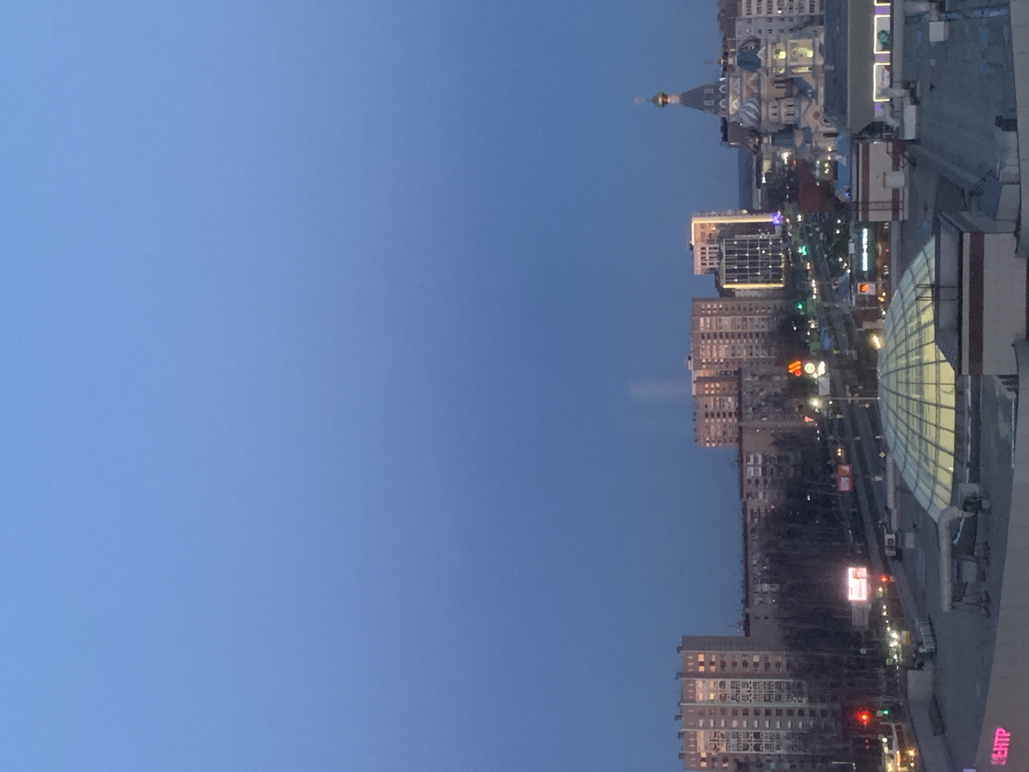 The image shows a partial cityscape of Izhevsk, Russia, at dusk. Dominating the right side are high-rise buildings, including a large shopping center with a glass roof. Further away, a road lined with trees and buildings stretches into the distance. In the center are more high-rise apartments. The top of an elaborate Orthodox cathedral is visible in the background. The sky is a clear, deep blue.