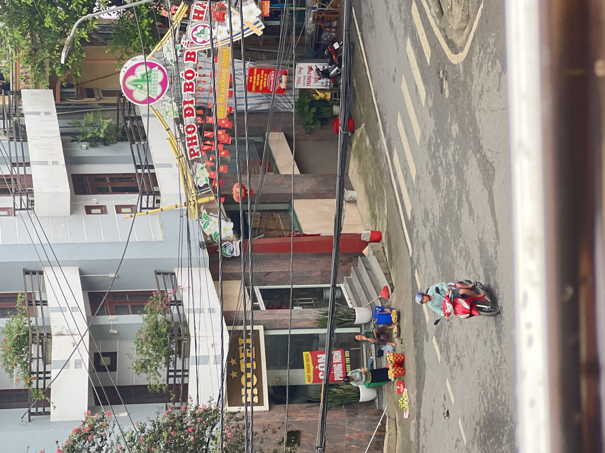 The image depicts a street scene in Bắc Hà, Vietnam. In the foreground, a man is riding a red scooter. The road has a pedestrian crossing. Behind the road, there is a building with a hotel sign that reads "HOTEL" with three stars. In front of the hotel, two women are sitting on the steps, and several baskets of fruits are placed on the ground. One woman appears to be tending to the fruits, while the other is seated. Above the hotel, a vibrant banner with Vietnamese writing and festive red flags suggests a local market or event. The architecture features balconies with potted plants, and numerous electrical wires crisscross the scene, characteristic of urban Vietnamese landscapes. The overall atmosphere suggests a busy street during daylight hours.