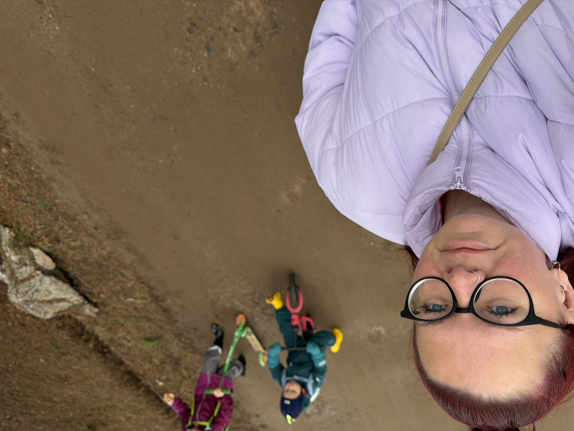 A low-angle, medium shot depicts a fair-skinned person with auburn hair wearing black-framed glasses and a light purple puffer jacket. They are looking down at the camera. Below them and to the left, a dirt path is visible, with a child riding a scooter and another child on a bicycle. This image was taken outdoors in Chaikovsky, Russia.