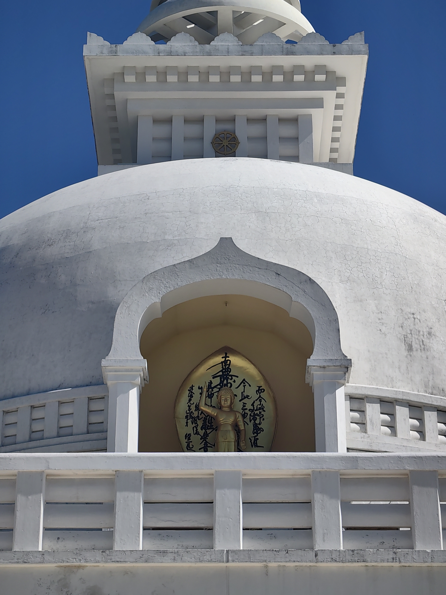 The image captures a close-up view of the upper section of a white stupa in Lumbini Bazaar, Nepal, under a clear blue sky. The stupa features a large, rounded dome with visible crack lines on its surface. Archways are carved into the side of the dome, revealing a circular golden disc inscribed with black calligraphy and a golden statue of a standing Buddha. Above the dome, there is a tiered, square structure with decorative architectural elements and a Dharmachakra symbol. A white, balustrade-like structure with horizontal openings runs across the foreground. The overall scene is static and focuses on the architectural details of the religious monument.