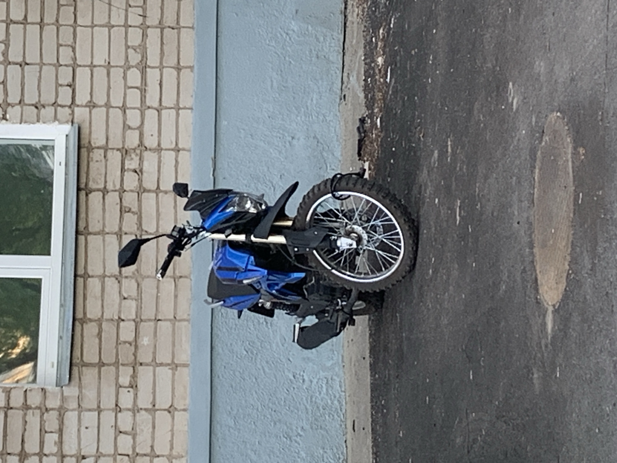 A blue and black dual-sport motorcycle is parked on a gray asphalt surface, possibly a sidewalk or parking area. The motorcycle is positioned next to a light blue wall that abuts a beige brick wall with a white-framed window. The surrounding environment suggests an urban area in Chaikovsky, Russia. There are no people visible in the image.