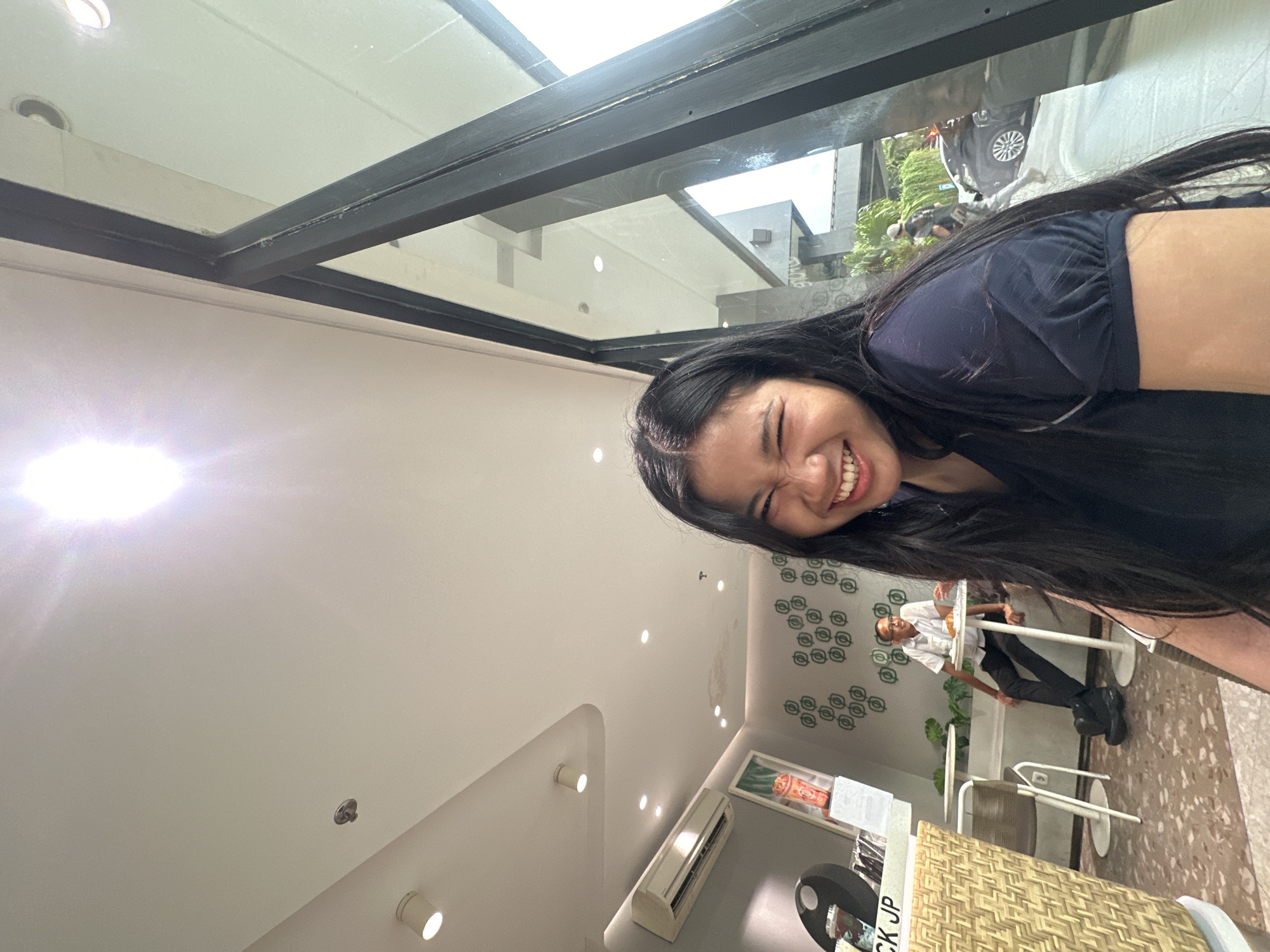 The image depicts a young woman with long, dark hair smiling broadly. She's positioned at the top right of the frame, with a glimpse of her dark blue top visible. The camera angle is from below, capturing the ceiling and exterior view.
The interior features a white ceiling with recessed lighting. An air conditioning unit is mounted on the wall. A person wearing glasses, a white shirt, and black pants is visible sitting at a table. The wall behind them has a pattern of circular designs.
The exterior is visible through a window with a black frame. Parts of buildings and a vehicle are seen, as well as some greenery. The scene appears to be in North Jakarta, Indonesia.