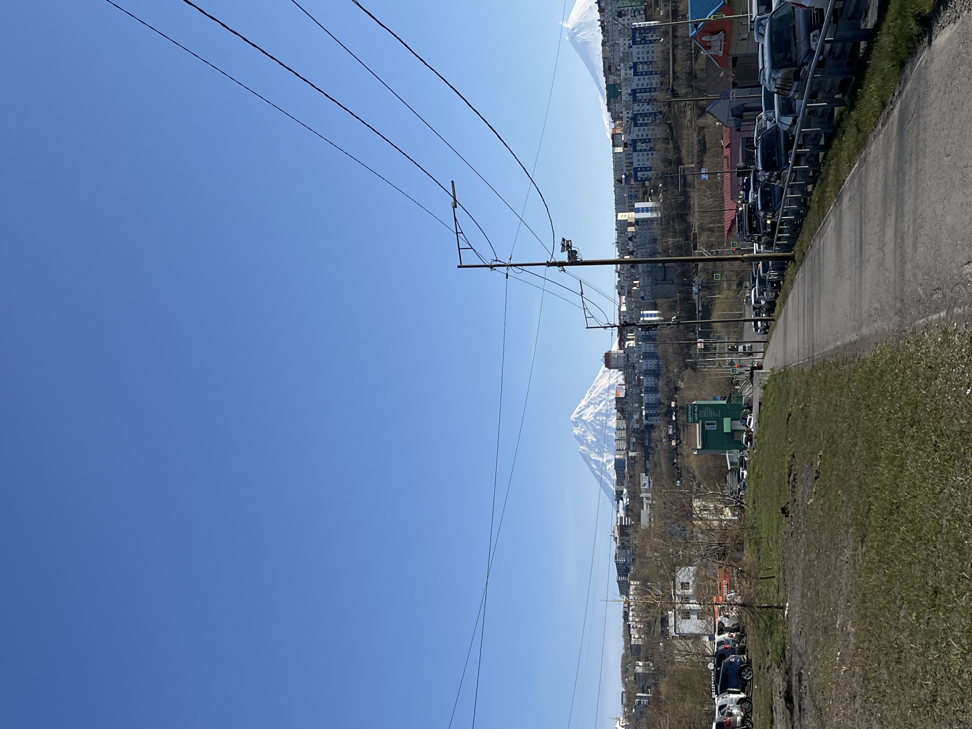 The image depicts a cityscape in Petropavlovsk-Kamchatsky, Russia. The scene is captured from a slightly elevated perspective, showcasing a view of the city against a clear blue sky. In the background, a snow-capped mountain is visible. Power lines stretch across the sky, adding a linear element to the composition. Buildings of varying heights and architectural styles make up the urban landscape. A road runs along the right side of the frame, with parked cars lining one side of it. A patch of grass is visible between the road and the buildings.