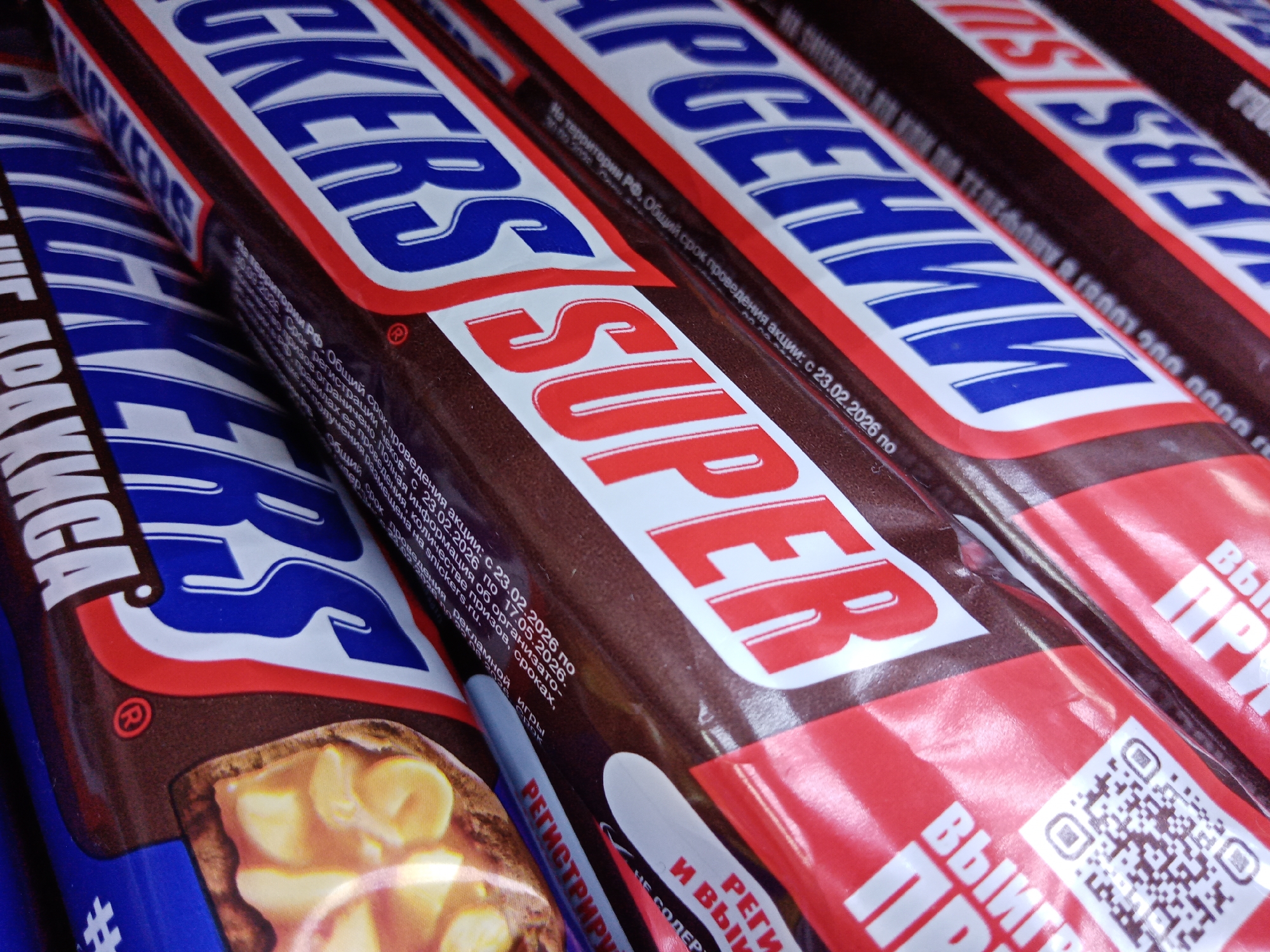 This close-up shot captures several Snickers chocolate bars arranged in a row, likely on a store shelf or display, in Elektrostal, Russia. The primary subjects are the packaged "Snickers Super" bars, identifiable by their prominent branding in red and white letters on a brown background, outlined with red. The bar on the far left partially shows a different Snickers variant, indicated by the Cyrillic word for "peanuts" ("АРАХИСА") on its blue packaging, and an image of the bar's interior filled with peanuts and caramel.
The wrappers feature extensive text in Cyrillic, indicating a promotional campaign. Key visible phrases include "SNICKERS SUPER" and parts of "ВЫИГР" (Win) and "РЕГИСТРИРУЙ" (Register), alongside "И ВЫИГР" (and Win). Detailed promotional information is also visible: "На территории РФ." (On the territory of the Russian Federation). The overall promotion period is stated as "с 23.02.2026 по 17.05.2026" (from February 23, 2026, to May 17, 2026), with "Срок регистрации чеков: с 23.02.2026 по 30.09.2026" (Period for registration of receipts: from February 23, 2026, to September 30, 2026). Further details are advertised to be available on "snickers.ru." A QR code is visible on one of the red-packaged bars, likely for participation in this promotion. Another partial phrase, "НЕ СОДЕР" (does not contain), is also seen. The image highlights the distinctive Snickers logo colors of brown, red, white, and blue, under clear, even lighting.