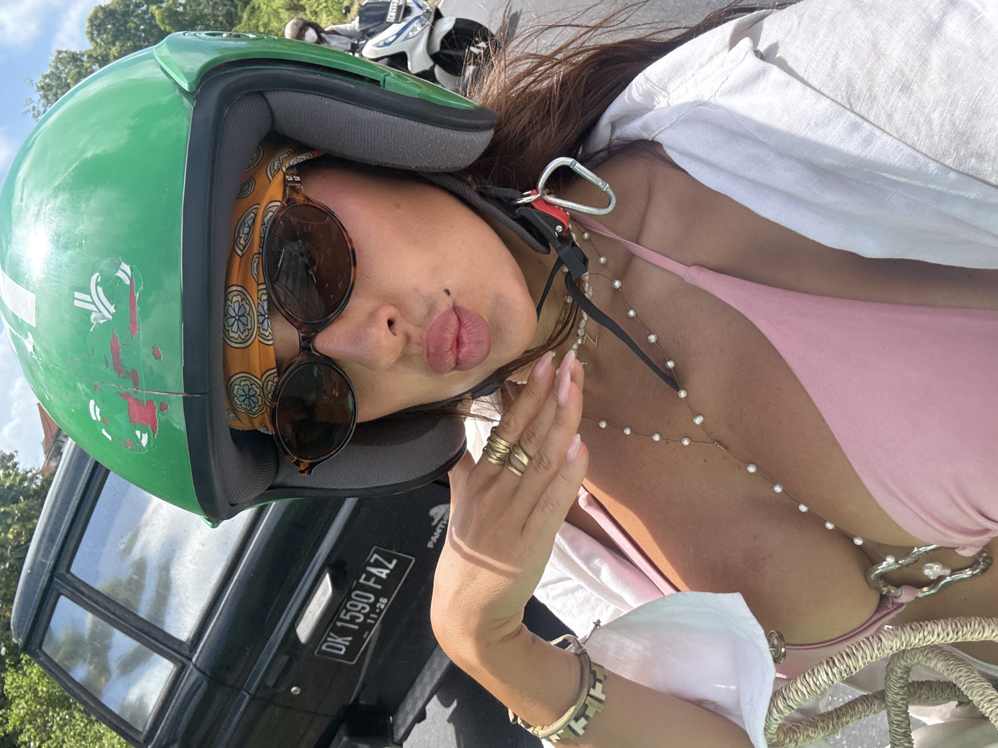 A selfie taken in Kuta Selatan, Indonesia, shows a woman wearing a green helmet and sunglasses. She has a patterned scarf under the helmet and is wearing multiple gold rings on her fingers. She has her lips pursed into a kiss. She is wearing a pink top and a white shirt, with a necklace visible on her chest. A black truck with a license plate reading "DK 1590 FAZ" is in the background.