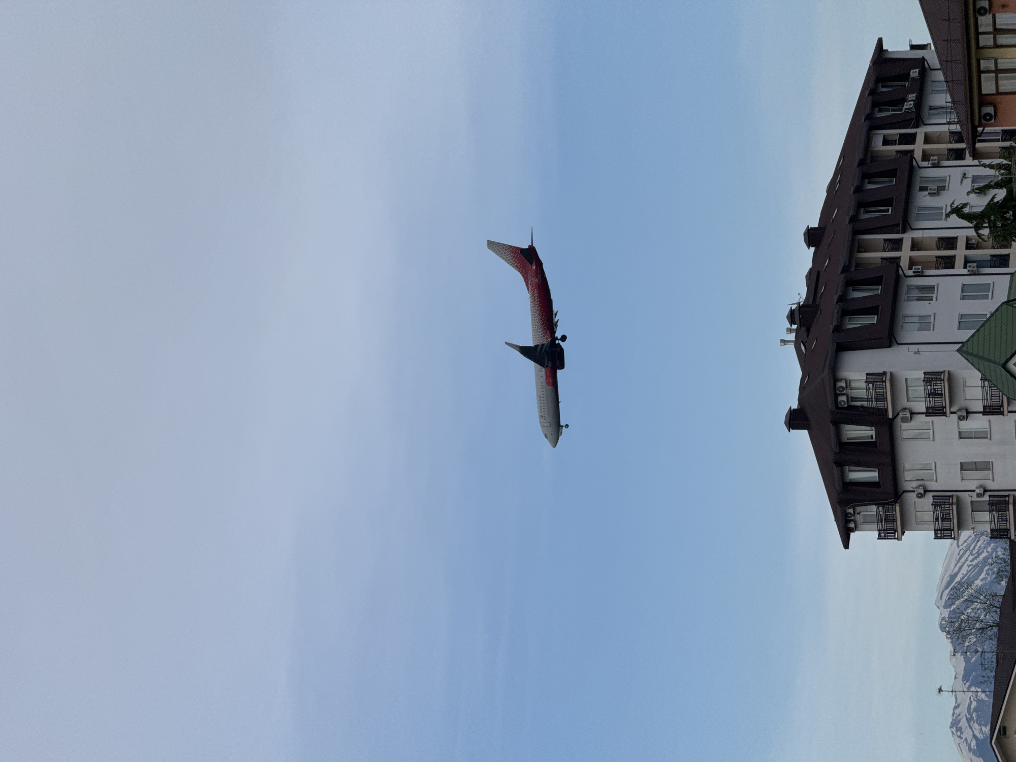 The image captures a paraglider soaring through the clear blue sky above Sochi, Russia. The paraglider, primarily red and white, is the focal point, positioned centrally in the frame. Adjacent to the paraglider is a tall, multi-story building with white walls and a dark brown roof. The building has numerous windows and balconies. In the distance, snow-capped mountains are visible.