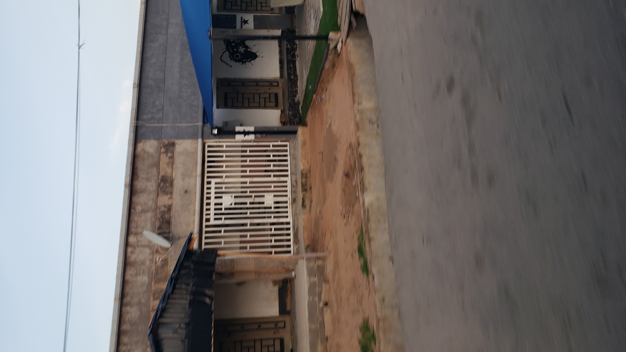 The image depicts a street scene in Jalingo, Nigeria, showing the facades of several buildings. On the left, a structure with a dark, corrugated metal roof extends over an entrance. Adjacent to it is a white metal gate, offering a view into a courtyard area. The gate is positioned in front of a building with a textured, weathered wall. To the right of the gate, another building features a white facade decorated with a black graphic resembling an abstract design or spray paint art, and a blue, angular roof structure. In front of this building, there is a patch of artificial green turf and a small wooden ramp or bridge. The foreground is dominated by a paved road, with a concrete curb separating it from the dirt and sand-covered ground in front of the buildings. Power lines are visible in the sky above the structures. The overall impression is of an urban environment, likely a residential or commercial area, in Jalingo.