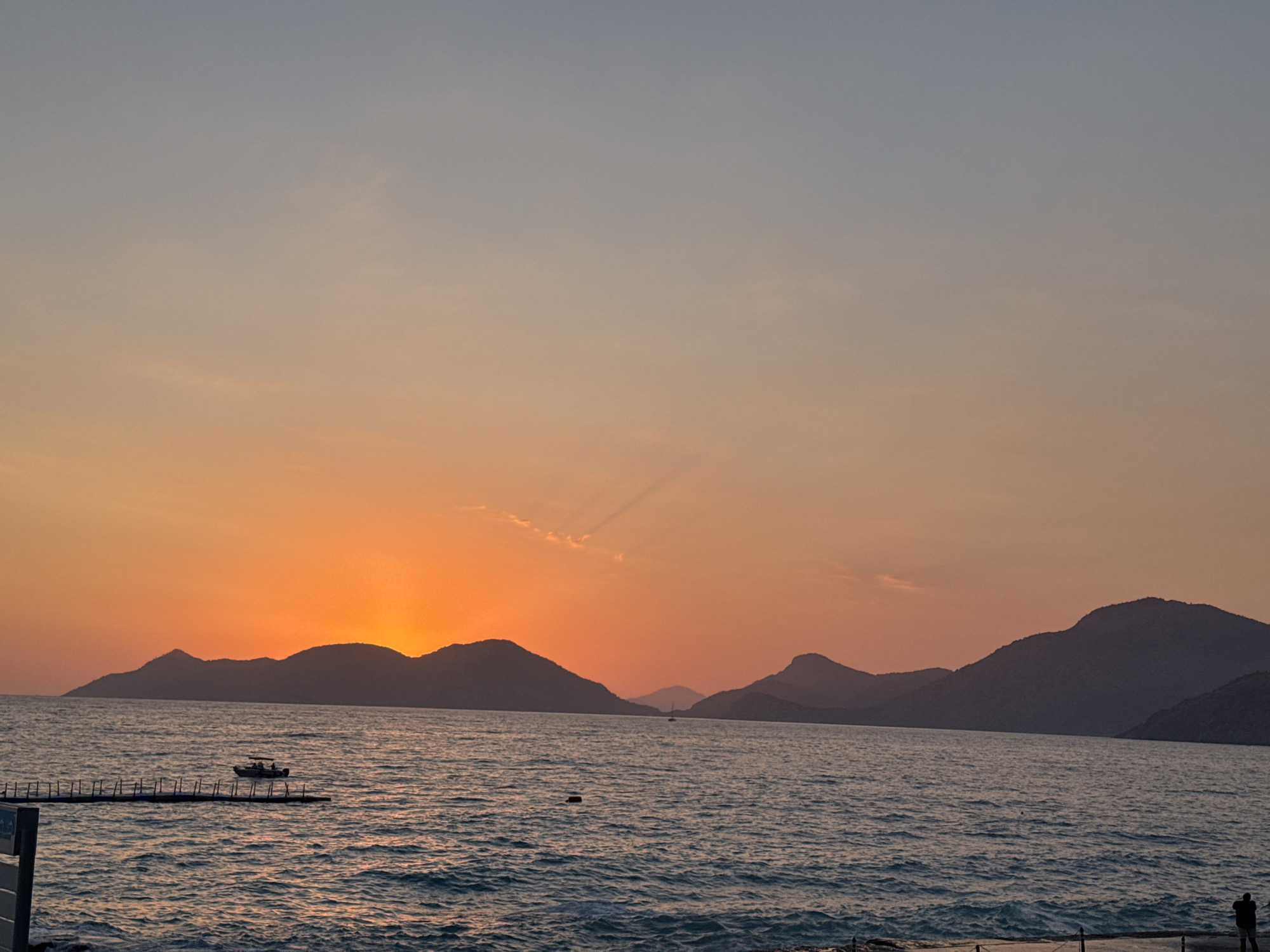 The image depicts a scenic sunset over the Aegean Sea in Fethiye, Turkey. Silhouetted against the vibrant orange and pink hues of the setting sun are a series of dark, mountainous islands stretching across the horizon. The sea is a deep blue, with gentle waves reflecting the sky's colors. In the foreground, a small motorboat with several occupants navigates the water, and to the left, a pontoon or pier extends into the sea. Further to the right, a solitary figure stands on a concrete walkway overlooking the water, suggesting a coastal promenade. The sky transitions from a warm orange at the horizon to a soft blue at the top, with faint wispy clouds present.
