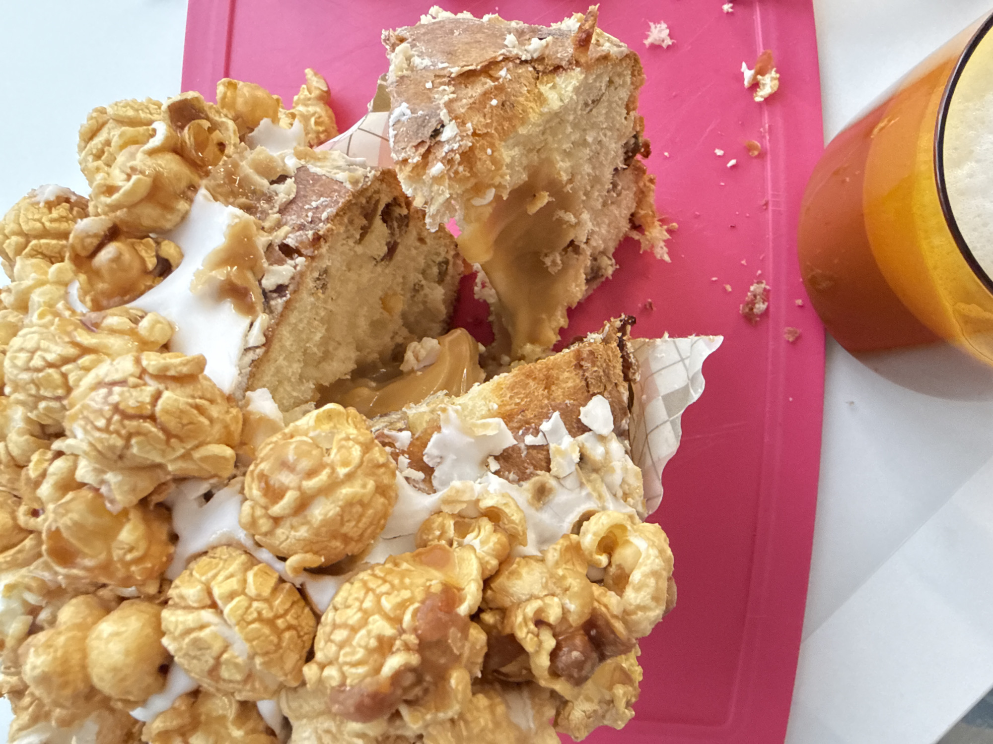 The image shows a partially eaten cake on a pink cutting board. The cake is decorated with caramel popcorn and white frosting and appears to have a caramel filling. To the right of the cake is a glass or pitcher containing a yellow-orange liquid, possibly juice or a smoothie. The background is a white surface. The scene does not provide any information to indicate it is located in Saint Petersburg, Russia.