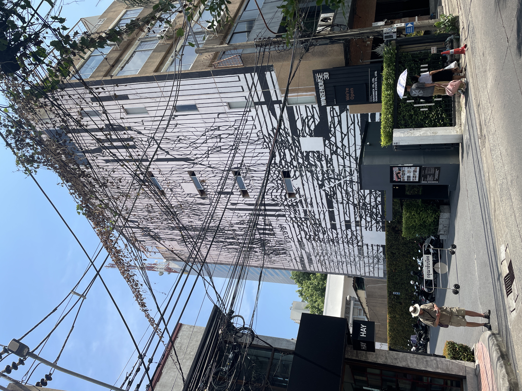 The image depicts a street scene in Bangkok, Thailand. An urban setting with a building featuring a modern, black-and-white facade is visible, adorned with abstract patterns. Tangled power lines dominate the foreground against a clear blue sky. A sign reads "Warehouse 30, We're Open," with the names "Chavanich Co., Ltd." and "Chavana Estate Co., Ltd." below.
On the sidewalk, a man wearing a hat and khaki-colored clothing is standing. Further down, a couple is walking under an umbrella near a hedge and a sign written in Thai. A motorcycle is parked along the sidewalk. The overall impression is of a busy urban area in Bangkok.