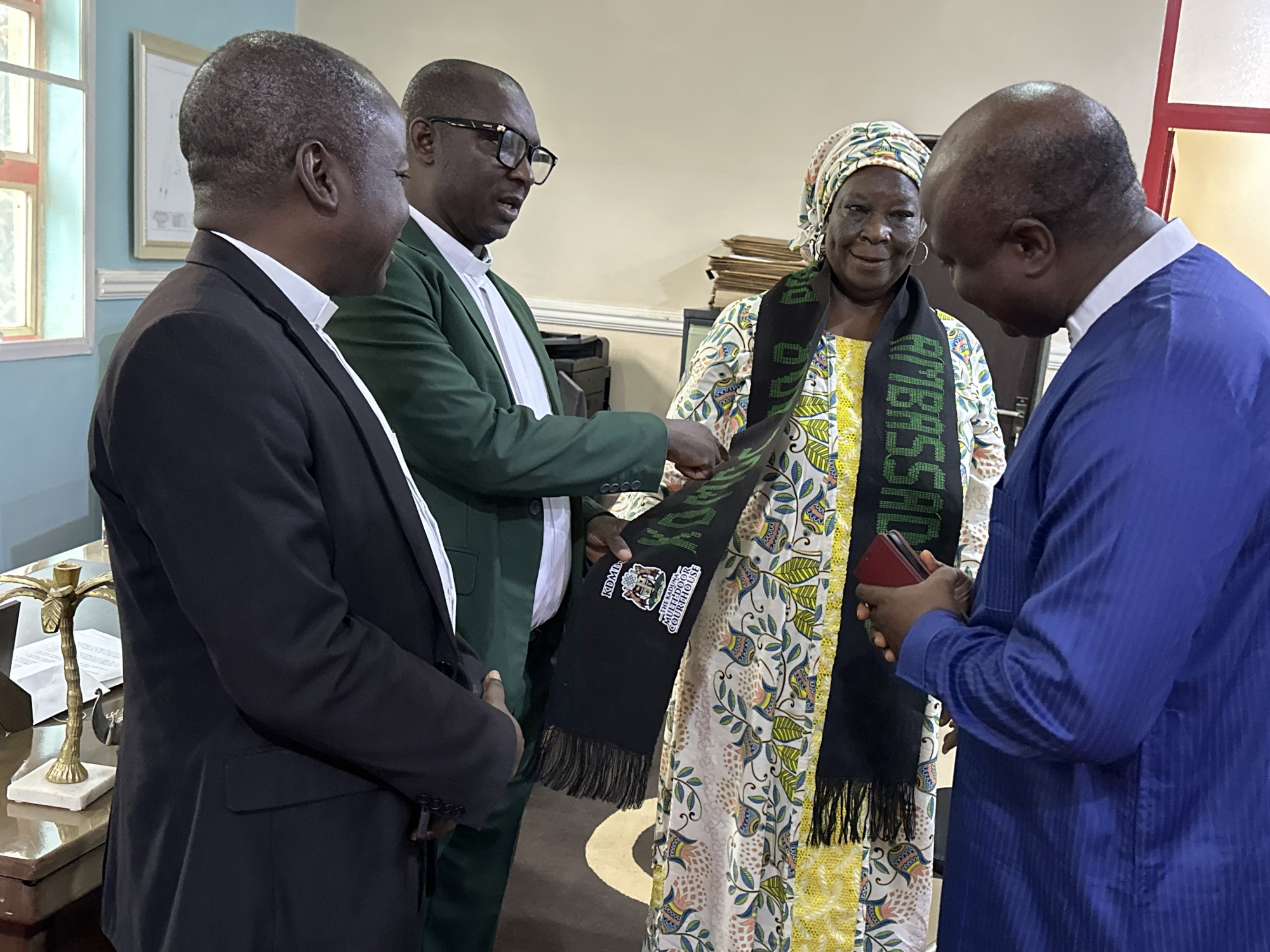 In a room with light-colored walls, four individuals are present. Two men in suits stand on the left, one in black and another in green. The man in the green suit appears to be speaking and gesturing towards a woman in the center who is wearing a headscarf and a patterned dress. She is holding a dark scarf with text and imagery, possibly a badge or crest. A second man, wearing a blue traditional garment, stands on the right, looking down at his hands which hold a red object, possibly a phone. The room appears to be an office or meeting space, with a desk visible on the left featuring a decorative palm tree sculpture. The overall lighting suggests an indoor setting during the day.