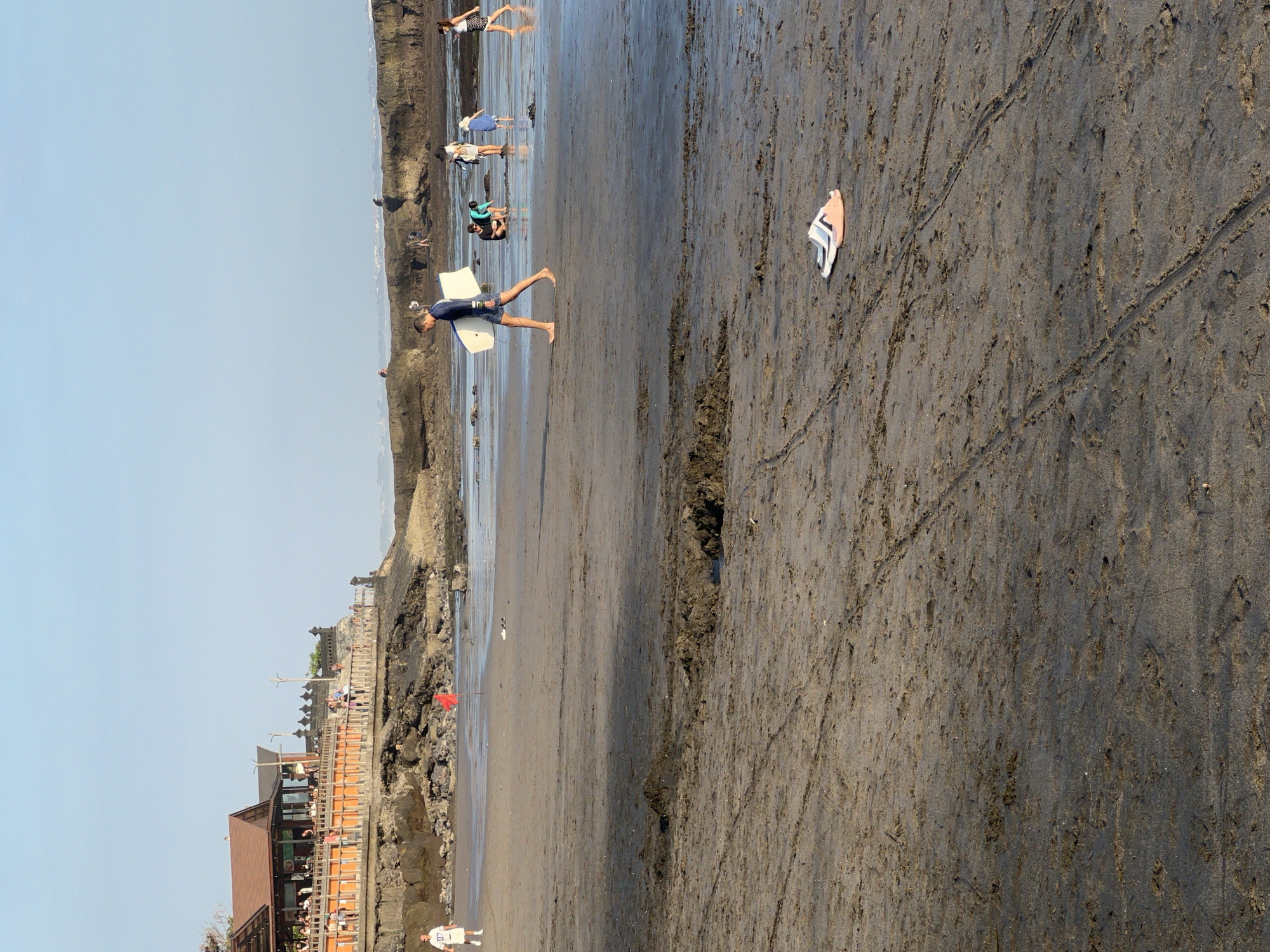 The image captures a beach scene in North Kuta, Indonesia, with a building and a promenade in the background. The foreground is dominated by wet sand with tire tracks and puddles. In the mid-ground, people are scattered across the beach. A man in a blue shirt and shorts walks towards the left, carrying a white surfboard. To his right, a group of people are gathered near the water's edge. Further to the left, a person in a white shirt and shorts stands near a red flag. A pair of flip-flops lie on the sand in the right mid-ground. The sky is clear and blue.