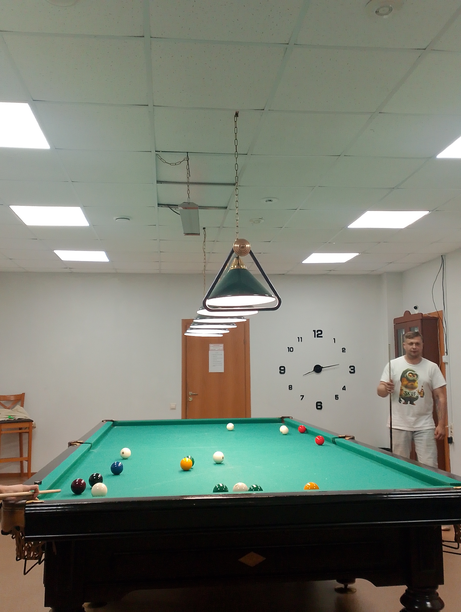 The image depicts an indoor billiard room. In the foreground, a dark-colored billiard table with a green felt surface is visible. Several billiard balls are scattered across the table, including white, red, blue, yellow, and green balls. One ball appears to be the cue ball. A person's hand is partially visible on the left side of the table, holding a cue stick, as if about to strike a ball. On the right side of the image, a man is standing near the billiard table, holding a cue stick. He is wearing a white t-shirt with a cartoon graphic and light-colored shorts. Behind the man, on the wall, is a large, modern clock with numbers. The room has a white ceiling with fluorescent light panels. A wooden door is visible on the left side of the image, and a cabinet is on the right. The overall impression is of a recreational space. The image was taken in Izhevsk, Russia.