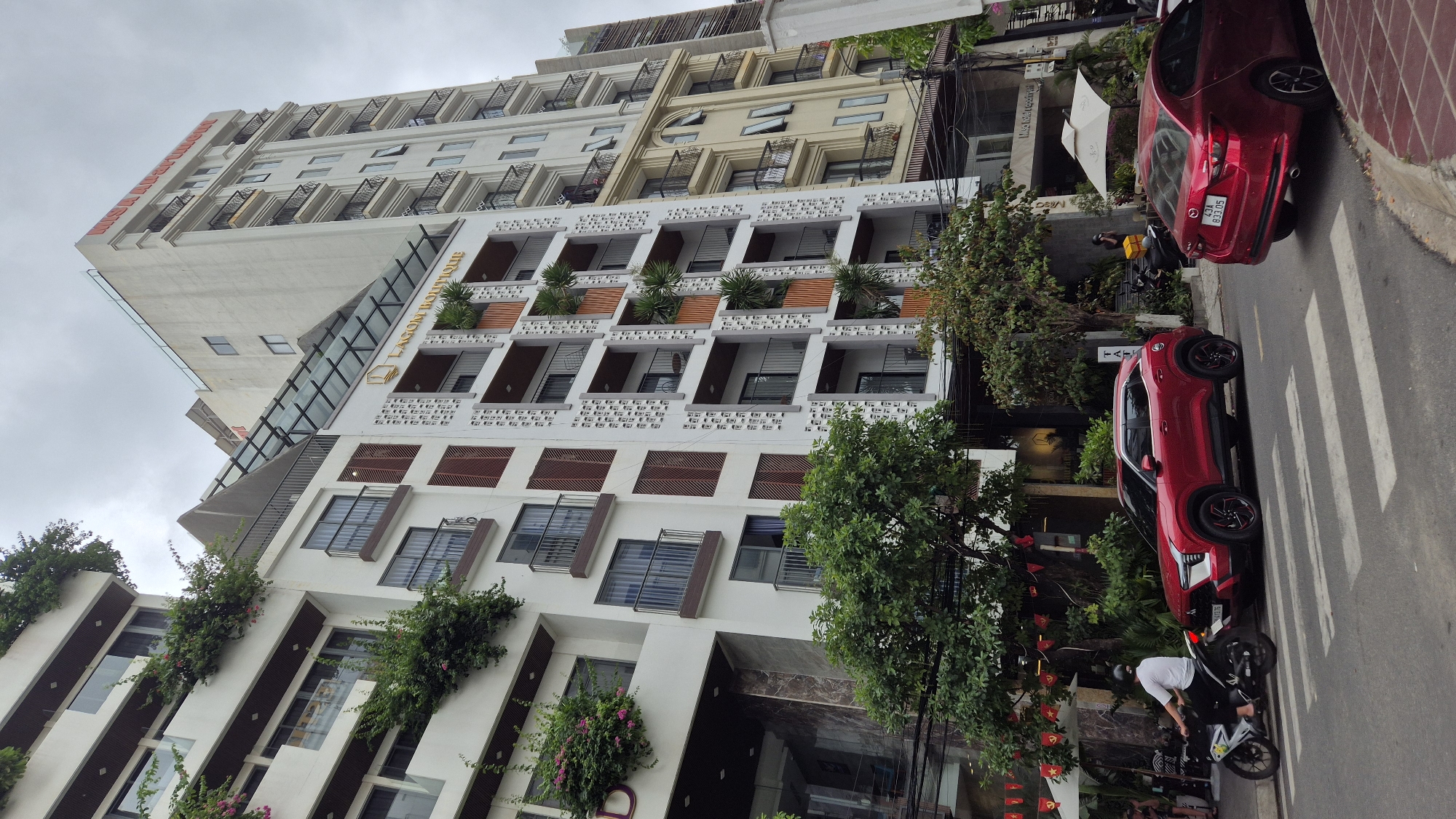 The image captures a street scene in Đà Nẵng, Vietnam, featuring modern multi-story buildings, a red SUV, a motorcycle, and lush greenery. The buildings are white with numerous balconies and decorative elements, including planters with plants and wooden shutters. One building has a sign that reads "LAGOM BOUTIQUE". The red SUV is parked on the street, and a person on a motorcycle is riding past it. Pedestrian crossings are visible on the road. The sky is overcast. The overall impression is of a well-maintained urban environment with a focus on architectural aesthetics and greenery.