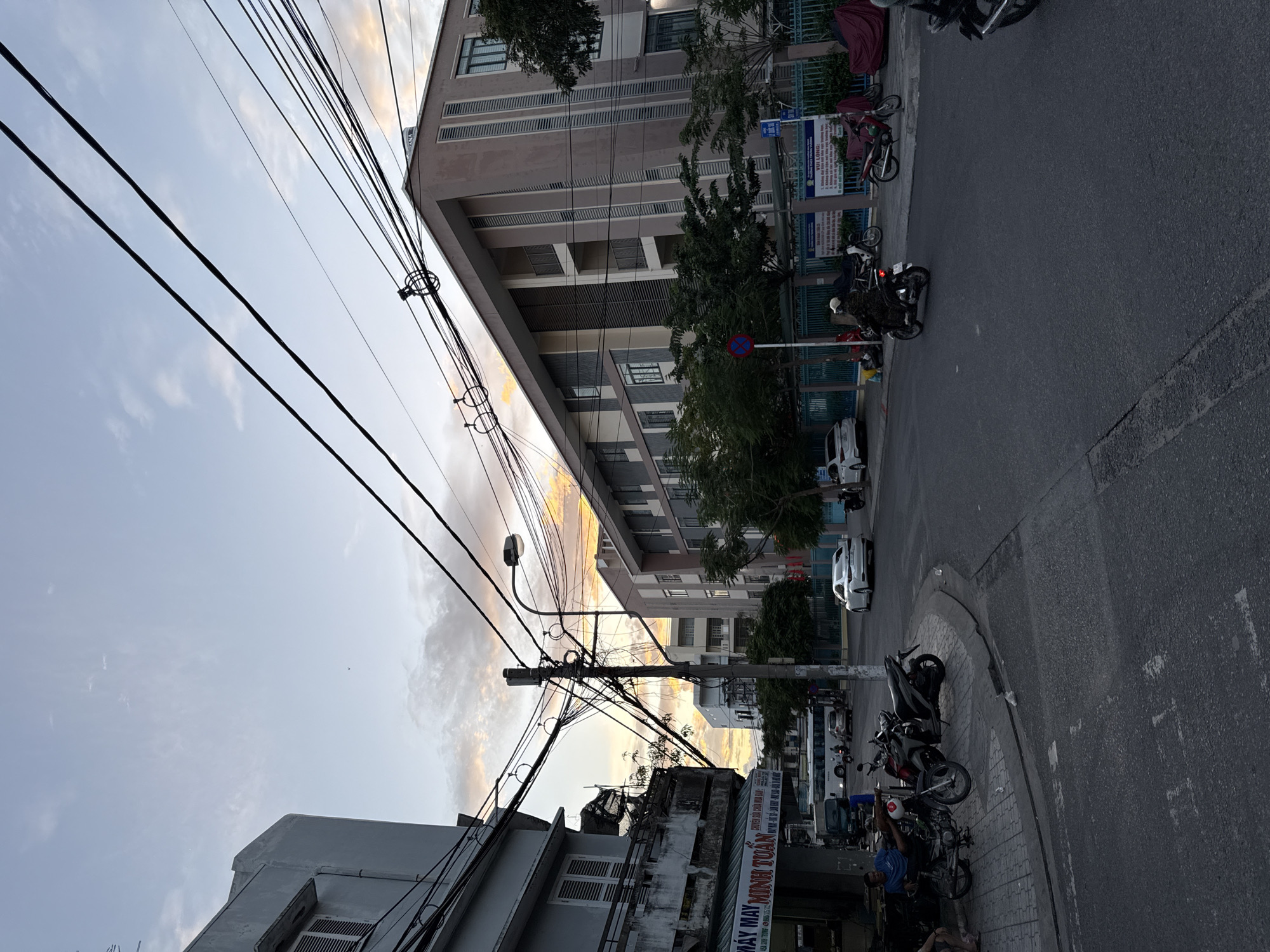 This is a medium shot, street-level, eye-level photograph taken in Nha Trang, Vietnam. The image captures a street scene at dusk, with a warm orange and yellow sunset sky visible in the background, peeking through a dense network of overhead power lines.
On the left side of the image, a weathered, multi-story building with a balcony and shutters is visible. A shopfront with a sign in Vietnamese, "MÁY MAY MINH TUẤN," is situated at its base. A few motorcycles are parked along the sidewalk in front of this building, and a person is seated on one of them.
The street curves into the distance, where more buildings and trees can be seen. A modern, multi-story building with a muted pink facade dominates the right side of the frame, its architecture characterized by repeating window patterns and vertical accents. A traffic light is visible on a pole in the middle ground.
Several motorcycles are in motion on the street. One motorcycle with a rider is heading away from the camera, while another is moving across the intersection. Cars are also present, including a white sedan driving away from the camera and another parked in the background. The street is paved and shows signs of wear.
The overall impression is of a busy urban environment during the transition from day to night, with a striking contrast between the utilitarian infrastructure of power lines and the natural beauty of the sunset.
