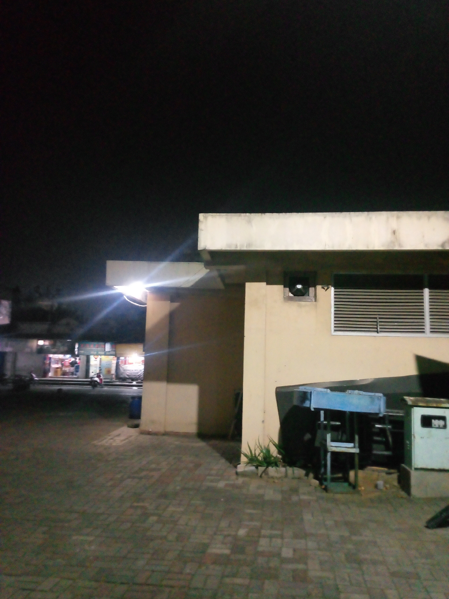 The image depicts an outdoor scene at night, likely in a commercial area of Bitung Jaya, Indonesia. A light-colored building with a flat roof and beige walls dominates the right side of the frame. To the left of the building, a bright light fixture emits lens flares, illuminating the overhead canopy and casting shadows.
In the background, several small businesses are visible, with illuminated storefronts and signs. A red and white motorcycle is parked in front of one of the shops. The ground is paved with interlocking brickwork.
To the right of the main building, a dark blue metal structure, possibly a food cart or service counter, is situated. In front of it, a small, green electrical box with a white label is present. A few plants are growing at the base of the building. The sky above is dark and appears cloudless.