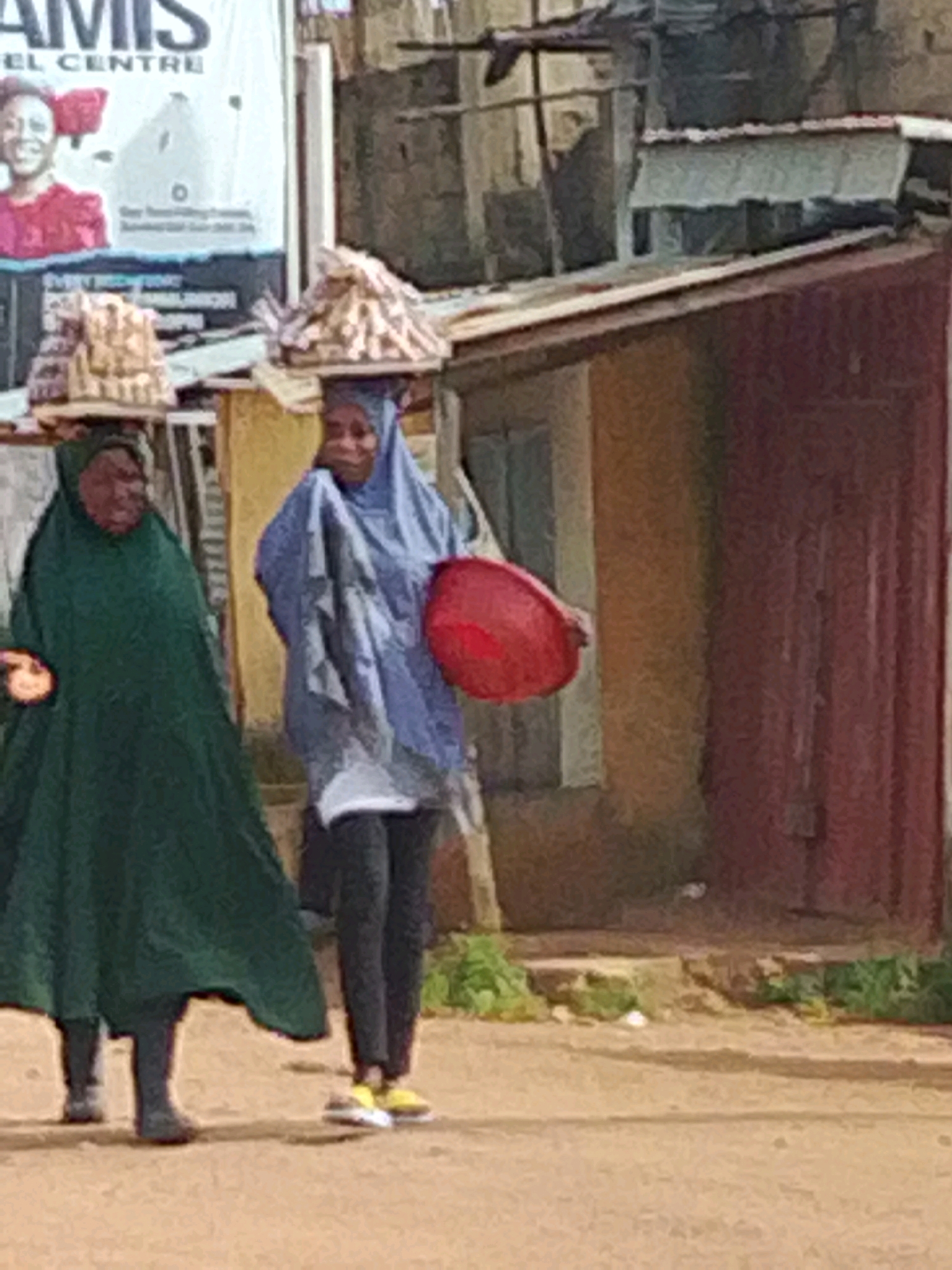 Two women are walking side-by-side on a dirt road in Jos, Nigeria. The woman on the left is wearing a dark green abaya and headscarf, carrying a tray of goods balanced on her head. The woman on the right is wearing a light blue abaya and headscarf, carrying a red bowl in her right hand and a tray of goods on her head. Behind them, a building with a red door is visible, and a large sign with text is mounted on a pole. The ground is dusty and uneven, with some patches of green vegetation.
