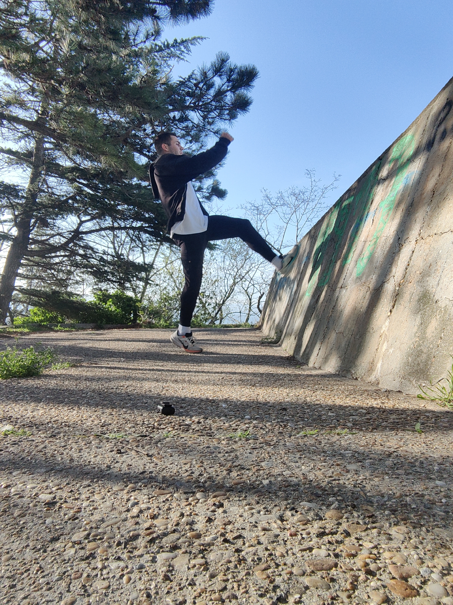 A dynamic, low-angle photograph captures a young man in mid-action outdoors in what appears to be a park or urban green space in Varna, Bulgaria. He is seen in profile, airborne, with his right leg extended high against a weathered concrete wall, and his arms raised, suggesting a parkour move, kick, or athletic stretch. He wears a black hoodie with a white section, black athletic pants, and athletic shoes with white socks. His expression is focused and determined.
The setting is characterized by a rough gravel path in the foreground, illuminated by strong sunlight that casts long shadows across the ground and wall. To the left, a large, bushy evergreen tree dominates the upper frame, its dark needles contrasting with the clear, bright blue sky. In the mid-ground, behind the man and the wall, the bare branches of deciduous trees are visible against the sky. The concrete wall, serving as a backdrop for the man's action, is textured and features faded green graffiti. On the gravel path in the foreground, a small, dark object, possibly an action camera, rests on the ground. The bright lighting and clear sky suggest it is daytime, likely morning or late afternoon.
