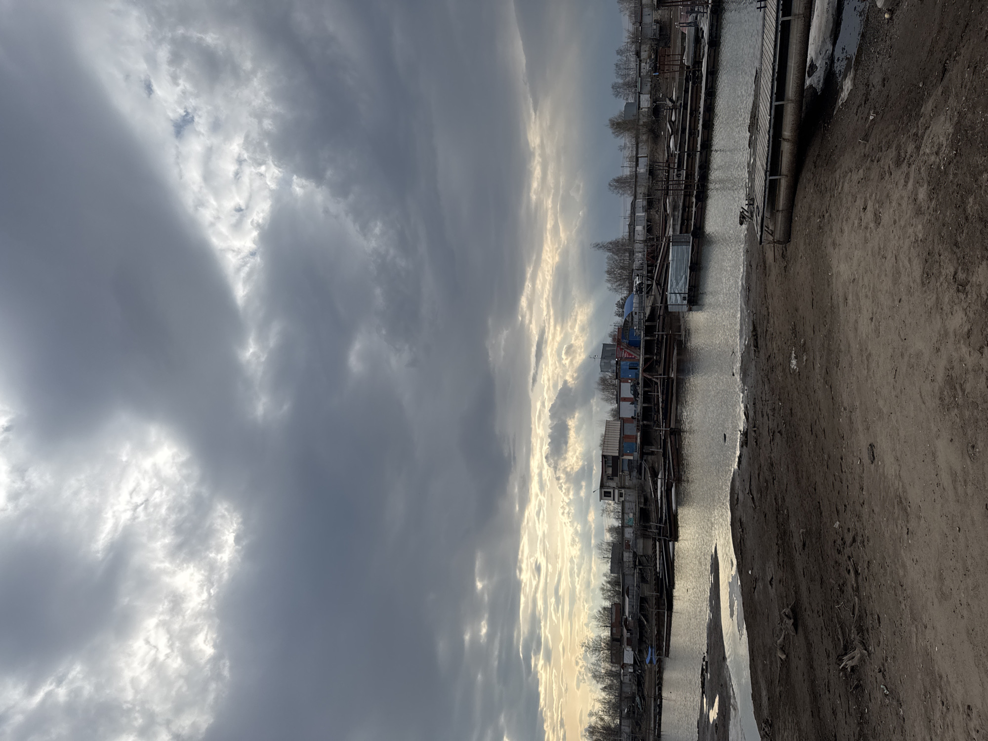 The image depicts a waterfront scene in Chaikovsky, Russia under a dramatic sky. Heavy, gray clouds dominate the upper portion of the frame, with patches of bright, filtered sunlight breaking through, illuminating the horizon. The lower portion shows a body of water, likely a river or channel, reflecting the sky. Along the waterfront, there are a series of small structures and boathouses, appearing somewhat rundown, lining the water's edge. A dirt or gravel bank runs along the right side of the image, suggesting a path or road beside the water. No people are visible. The scene appears bleak and industrial.
