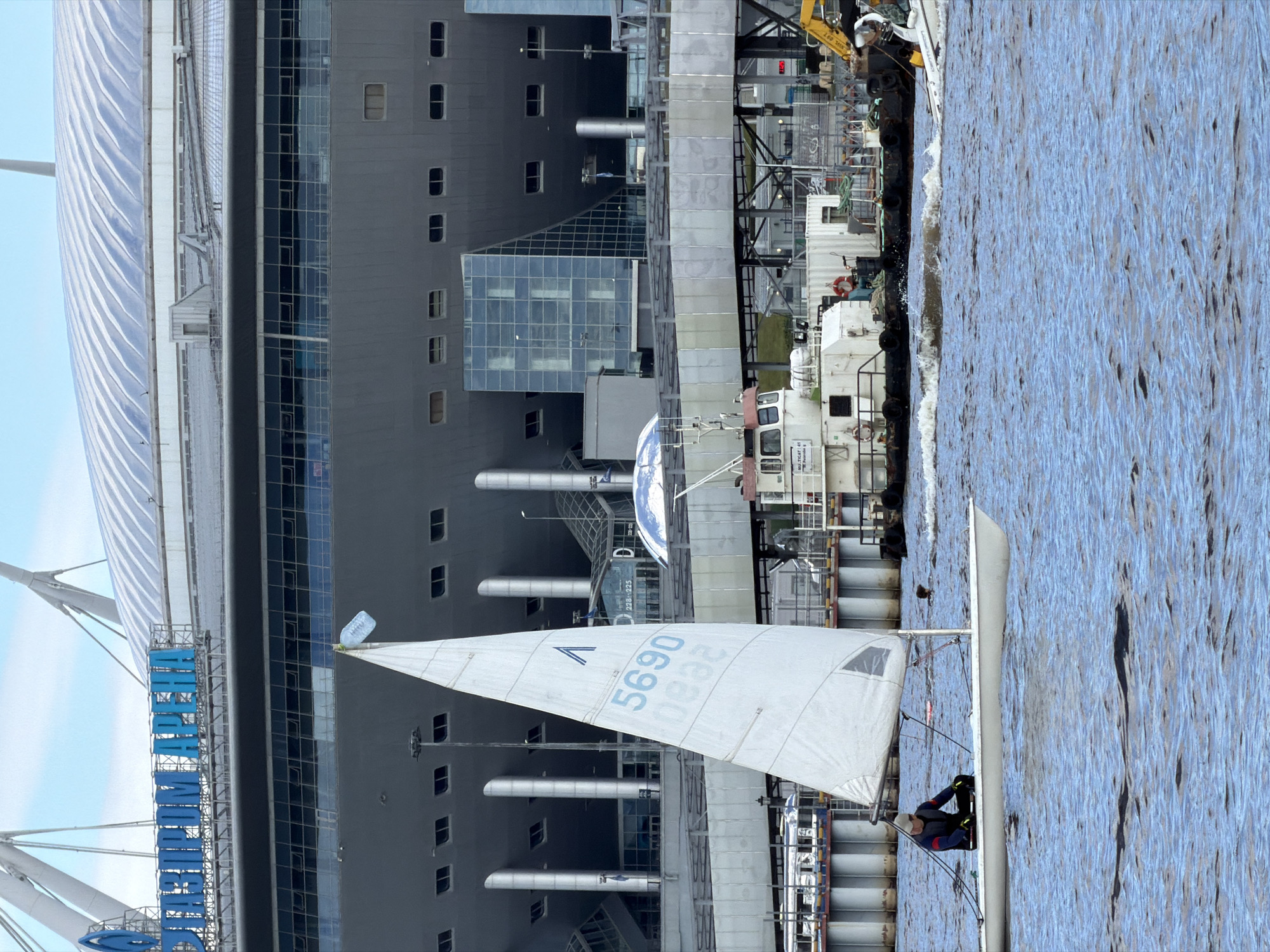 The image shows a body of water with a small sailboat in the foreground, on which a person is seated. The sailboat has a white sail with blue markings that include the numbers "5690" and "0822". Behind the sailboat, a large stadium is visible, identified by blue lettering on its facade as "ГАЗПРОМ АРЕНА" (Gazprom Arena). Several boats and structures, including a tugboat and what appears to be part of a construction site, are positioned on the water behind the sailboat and in front of the stadium. The stadium is a modern, domed structure with a gray exterior and a light-colored, ribbed roof. The water is choppy and reflects the sky. The scene is likely set in Saint Petersburg, Russia, given the prominent "Gazprom Arena" which is a well-known landmark in the city.