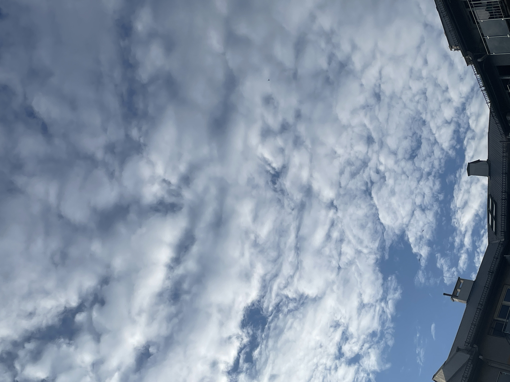 The image shows a view of the sky with many white clouds and some patches of blue sky visible between them. A small, dark speck, possibly a bird, is present in the sky. The upper right portion of the image shows the edge of a building with windows, a balcony, and a chimney-like structure. The location is unknown.