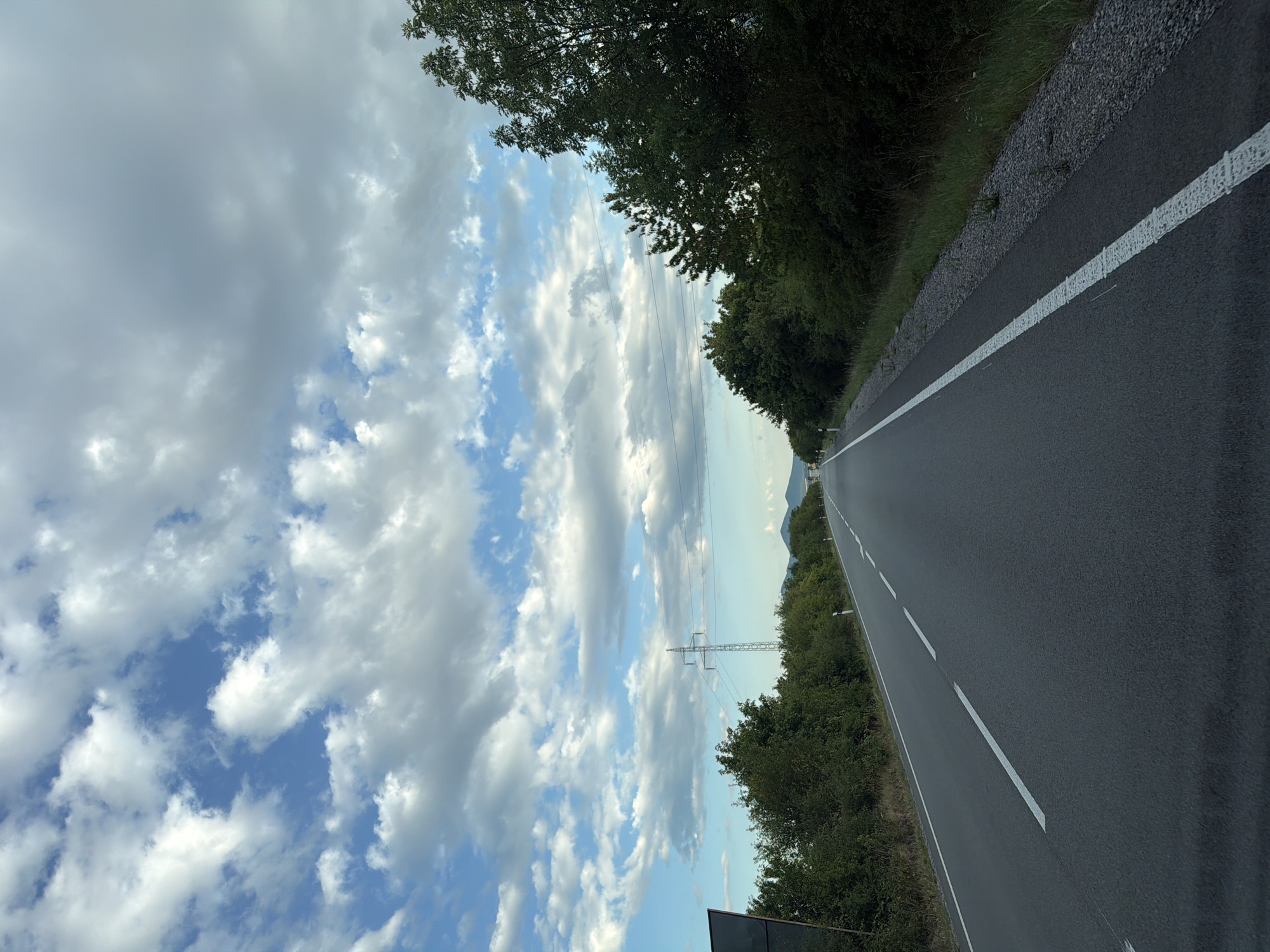 The image depicts a highway in the city of Luhyňa, Slovakia, under a partly cloudy sky. The road is paved and asphalt, with a white line marking the lane. Lush green trees and vegetation line both sides of the road. In the distance, a power line tower is visible. The perspective suggests the photo was taken from a vehicle moving along the highway.