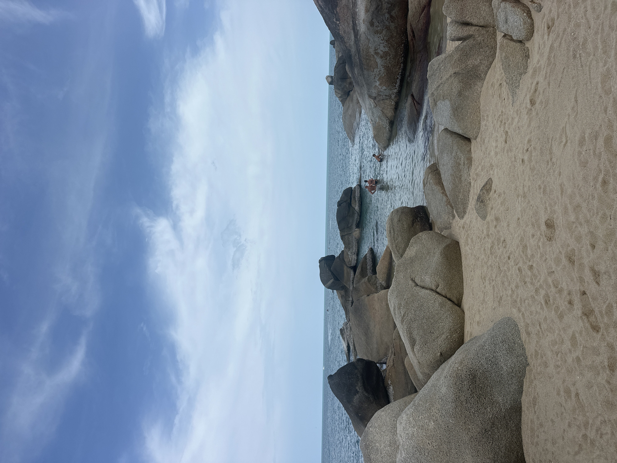 This is a wide-angle, eye-level, outdoor, vertical photograph of a sandy beach with large boulders and the ocean. In the foreground, a sandy beach stretches from the bottom of the frame. Several large, smooth, grey boulders are scattered on the sand and in the shallow water. In the middle ground, three people are visible in the clear water between the boulders. They appear to be swimming or wading. The ocean extends to the horizon line, where it meets a pale blue sky with scattered white clouds. The overall scene is serene and natural, evoking a sense of a tropical beach destination. The location is the coast of Ko Samui District, Thailand.
