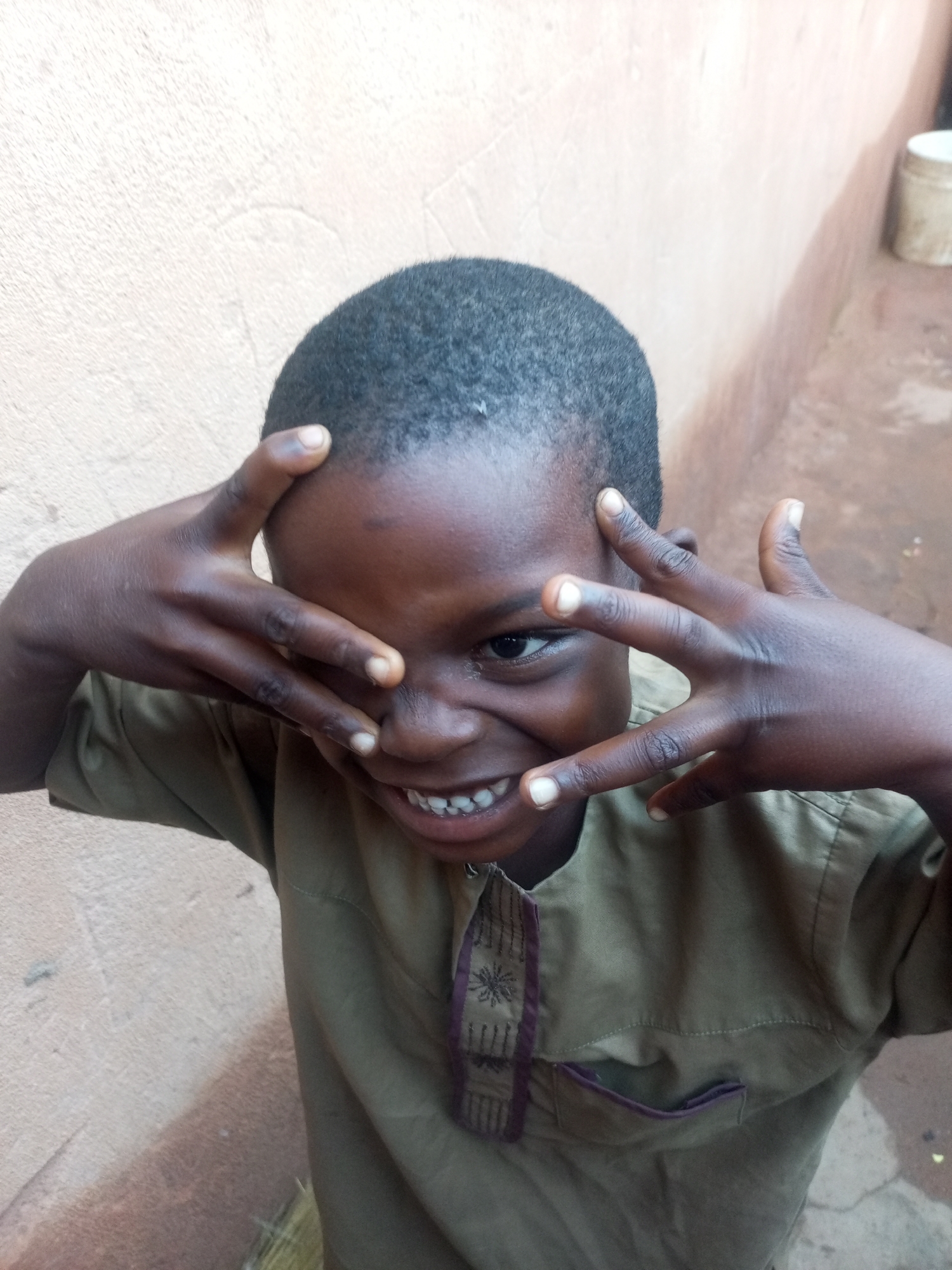 A young boy with dark skin and short, dark hair is shown from the chest up, looking towards the camera. He is wearing a khaki-colored shirt with purple trim. His hands are raised to his face, with his fingers spread and obscuring parts of his forehead and cheeks, and his mouth is open in a wide smile, revealing white teeth. He appears to be indoors or in a covered outdoor space, with a textured, light-colored wall behind him. To the right of the boy, a section of a reddish-brown wall is visible, and further to the right, a white bucket or container sits on the ground. The ground appears to be a dirt or concrete surface.