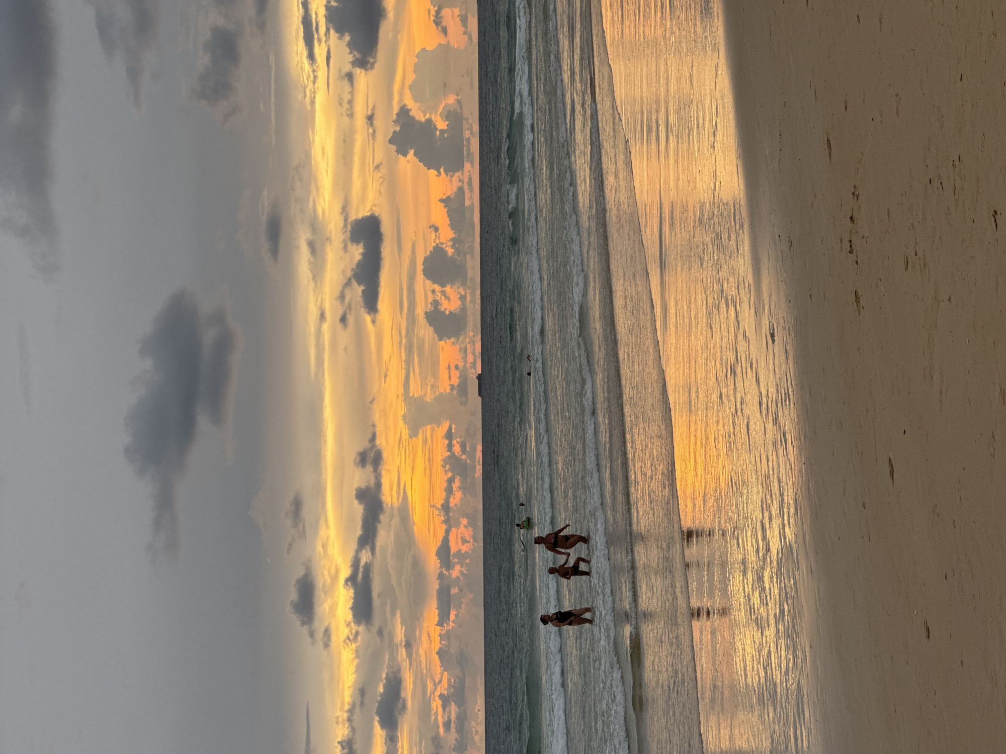 The image depicts a serene beach scene at sunset, with the sky ablaze with vibrant orange and yellow hues, interspersed with dramatic grey clouds. The sun's golden light casts long shadows across the wet sand and reflects brilliantly on the gentle waves as they lap the shore.
In the shallow water, three individuals are visible, enjoying the warm evening. Two appear to be adults, and one is smaller, possibly a child. They are standing or wading, their silhouettes dark against the illuminated sea. Their presence suggests a leisurely and peaceful moment, perhaps a family outing or friends spending time together.
The beach itself appears sandy, with some scattered debris or natural elements visible on the drier parts of the sand. The water is calm, with small, rippling waves indicating a tranquil ocean.
The overall atmosphere is one of peacefulness and natural beauty. The dramatic sunset combined with the calm ocean and the relaxed figures on the beach creates a picturesque and calming scene. The time of day is clearly sunset, as evidenced by the colors of the sky and the long shadows. The location is a beach, and given the context, it is Karon, Thailand. There is no visible text in the image.