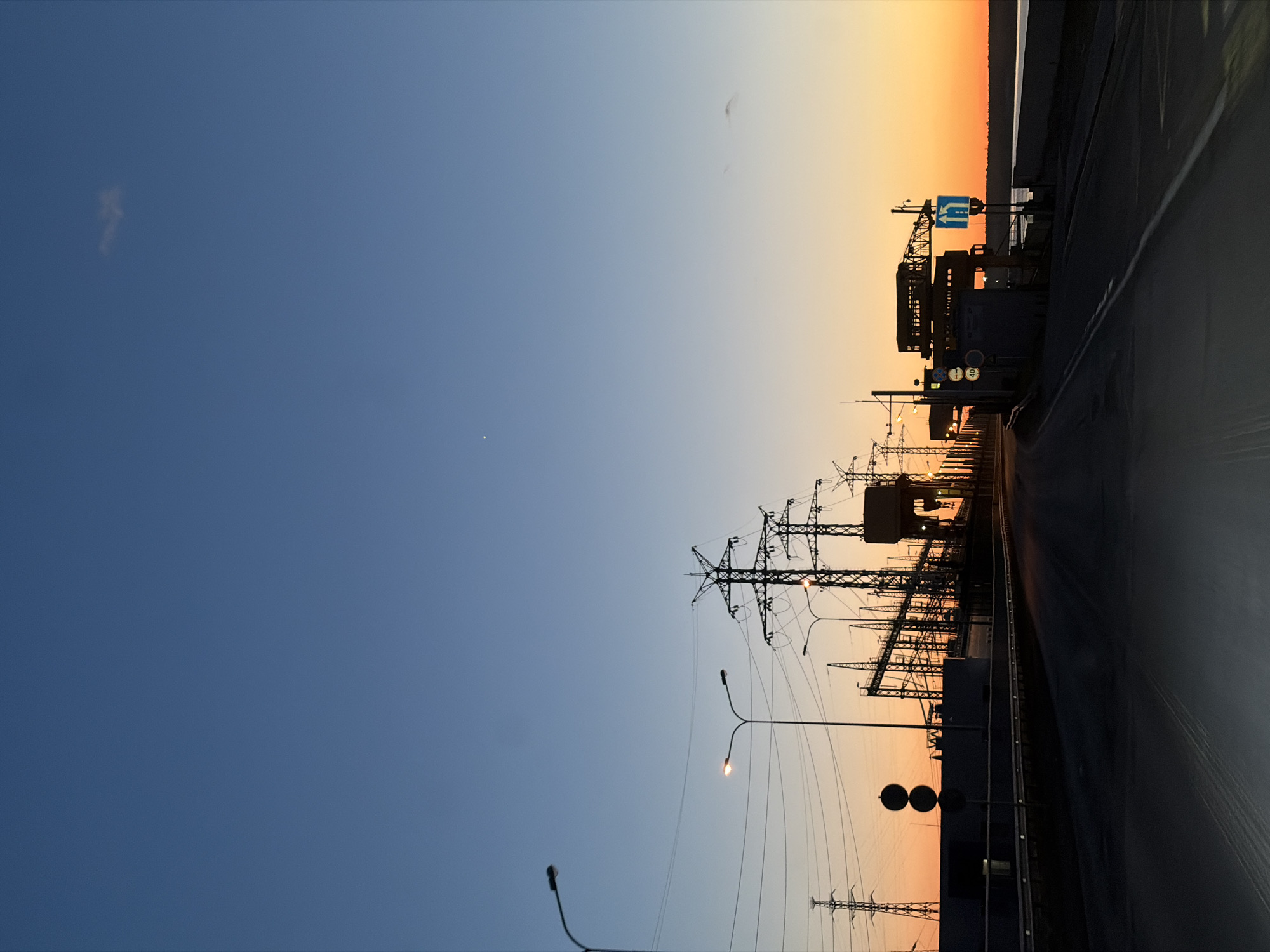The image shows a view from a roadway in Chaikovsky, Russia, at dusk or dawn. The sky transitions from blue to orange. An industrial area is visible to the right, with electricity pylons and power infrastructure silhouetted against the lighter sky. Street lights are present along the road. There is a road running alongside the industrial area, with a vehicle traveling on the road. A road sign is present in the top right area of the industrial area.