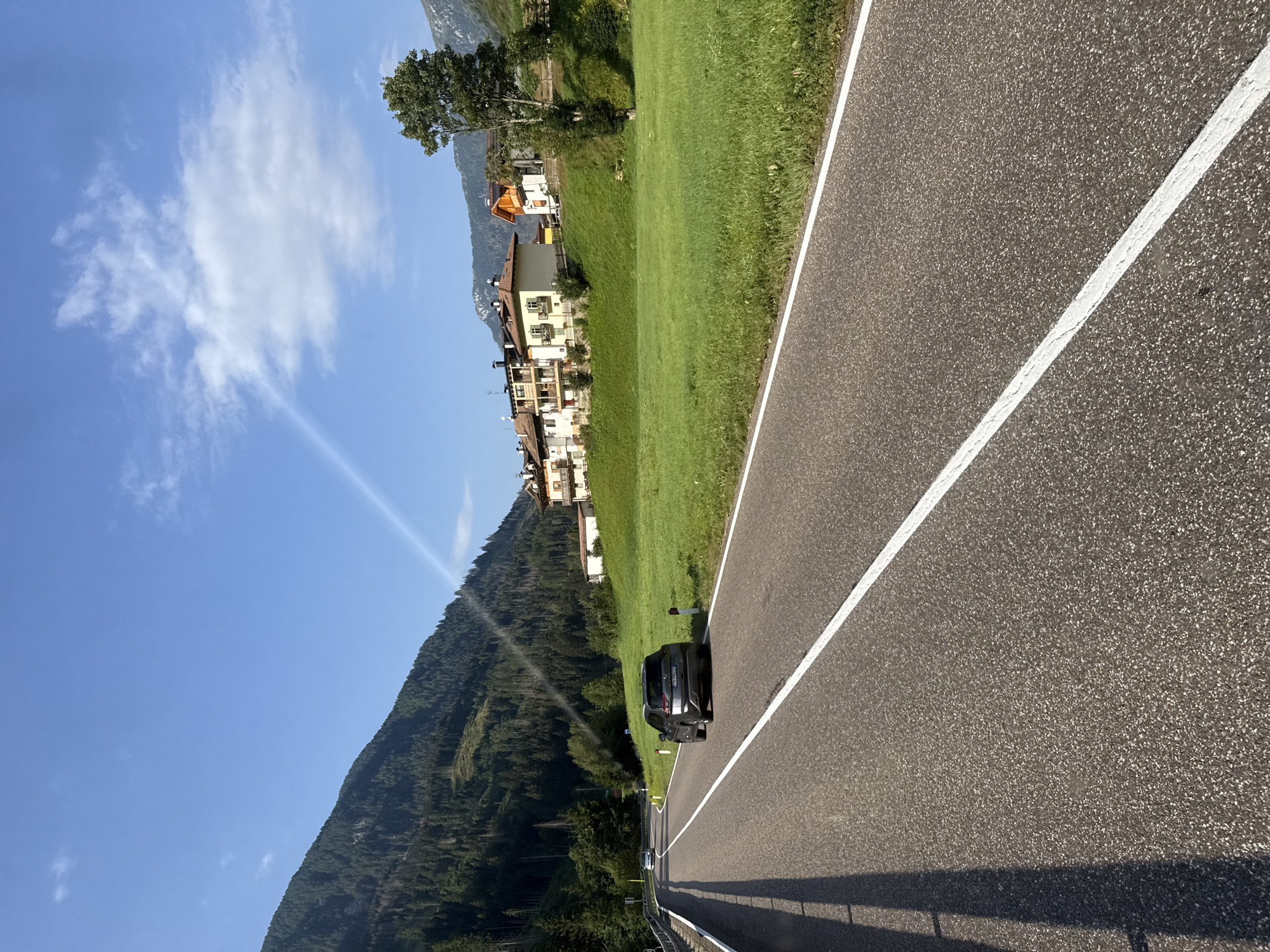 A scenic view captures a paved road winding through a mountainous region, possibly near Moena, Italy. The road is bordered by a vibrant green grassy field on the right, where several houses with tiled roofs are nestled. A dark-colored SUV is driving away from the camera on the road, and another car is visible further up the road. The left side of the road features a steep, tree-covered mountainside. The sky above is a clear blue with scattered white clouds, and a faint sunbeam is visible, suggesting a bright, sunny day.