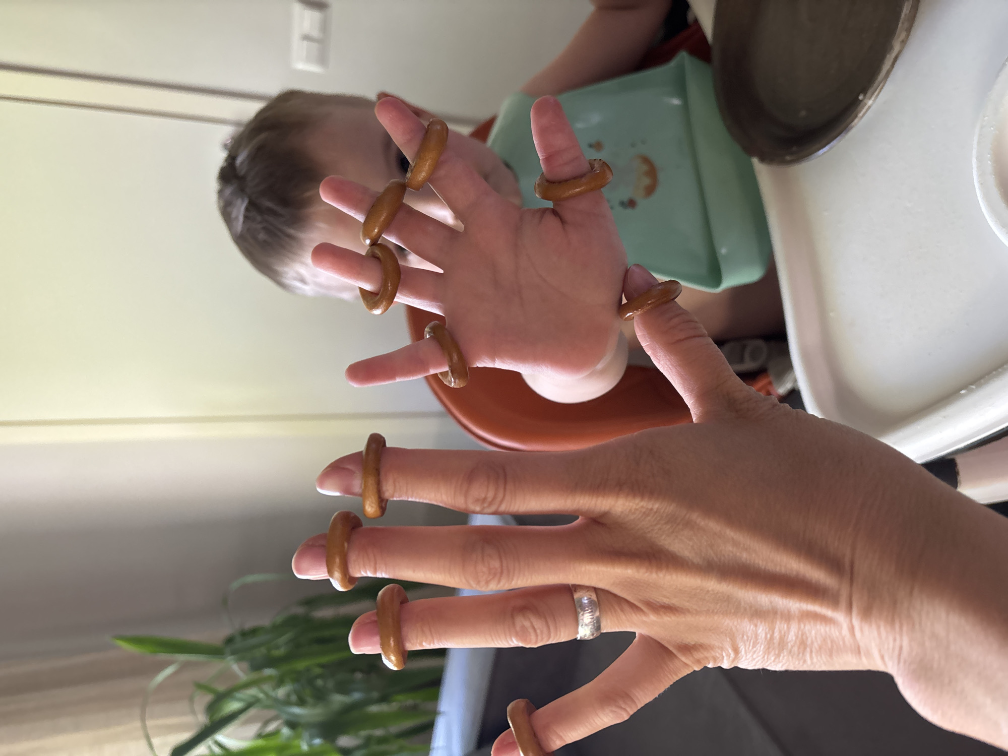 The scene shows two hands, one appearing to be that of an adult and the other belonging to a child, both adorned with small pretzel rings worn on each finger like jewelry. The adult hand features a silver ring on the ring finger. A child is seated in a highchair, wearing a green bib, in the background. A white tray table with dishes is also visible. The setting appears to be indoors, possibly a home. There's no evidence to suggest the image was taken in Chaikovsky, Russia.