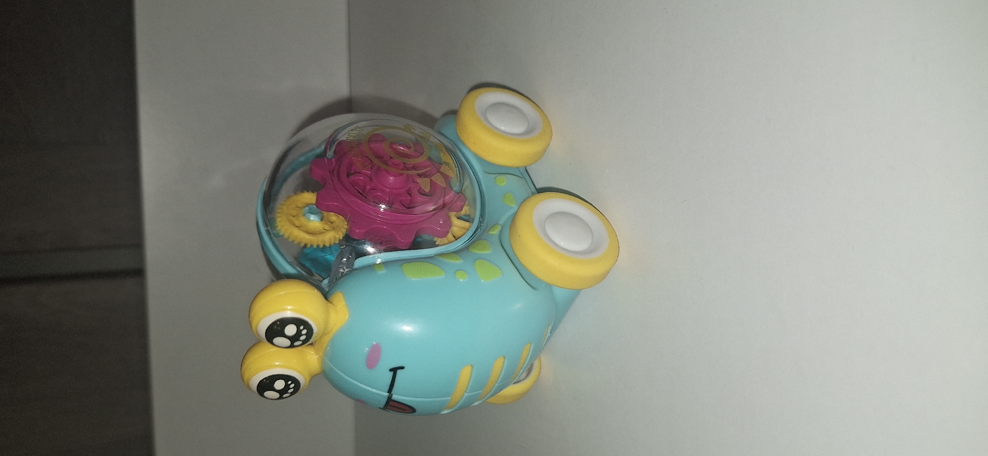 A close-up, vertically oriented shot showcases a blue toy snail with yellow wheels and a clear plastic dome on its back. Inside the dome, pink and yellow gears are visible. The snail has large, googly eyes with black pupils and white highlights. A small pink mouth with a red tongue is drawn on its face. The toy is placed on a light gray surface, possibly a wall or a table, with a dark gray area to the left, suggesting a corner or a contrasting background element. The lighting is diffused, and there are no indications of the time of day or weather. The setting appears to be indoors, likely a home or play area. There is no visible text in the image.