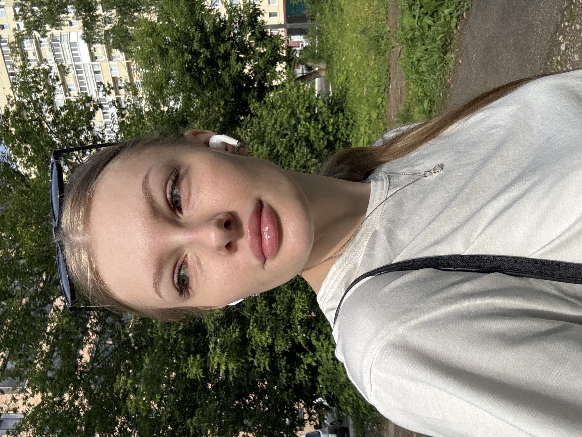 A young woman with blonde hair and fair skin is shown in a medium close-up, looking off to the left. She is wearing a white t-shirt, a silver necklace with a small pendant, and white wireless earbuds. Sunglasses are perched on top of her head. Her makeup includes prominent eyeliner and lip gloss. The background consists of lush green trees and a tall, multi-story apartment building. The setting appears to be an urban residential area, possibly in the city of Perm, Russia, given the architectural style of the building and the presence of trees common in Russian cities. The lighting suggests it is daytime with bright sunlight casting shadows. A paved path is visible in the lower right corner of the image.