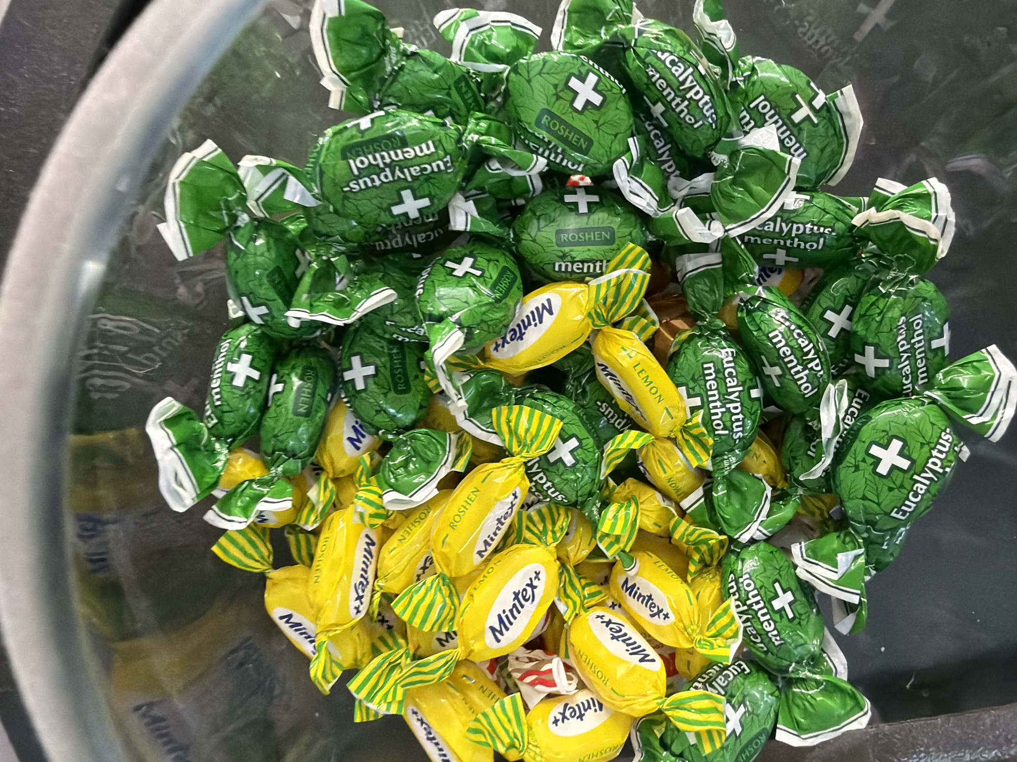 A pile of individually wrapped candies, some yellow and some green, are contained within a metal bowl. The yellow candies are inscribed with the brand name "Mintex+" and "Roshen" in white text, some also featuring "Lemon" in smaller text. The green candies are printed with "Eucalyptus menthol" and "Roshen" in white text, alongside a white cross symbol. The scene appears to be an indoor setting. The image was taken in Kyiv, Ukraine.