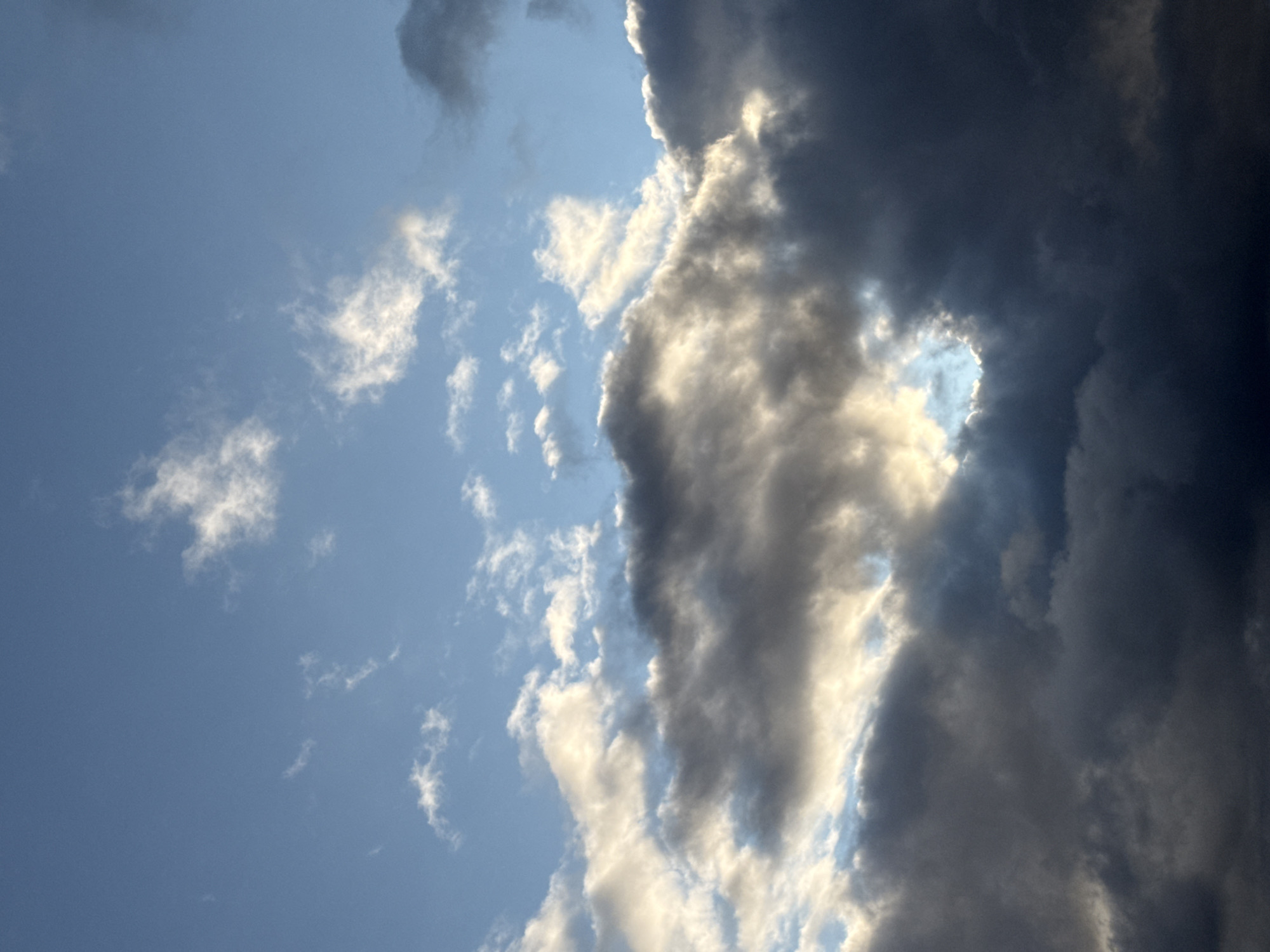 The image depicts a sky with a mix of light and dark clouds. The sky is a bright blue, with scattered wispy white clouds at the top. Towards the middle and bottom of the frame, darker, more ominous clouds are present, with shafts of sunlight breaking through in places, creating a dramatic effect. The lighting suggests it could be late afternoon or early morning. There are no people, subjects, or objects in the image, nor any discernible urban features, indicating it is a landscape or skyscape photo. It is impossible to determine a specific location within the city of Uuesalu, Estonia based solely on this image.