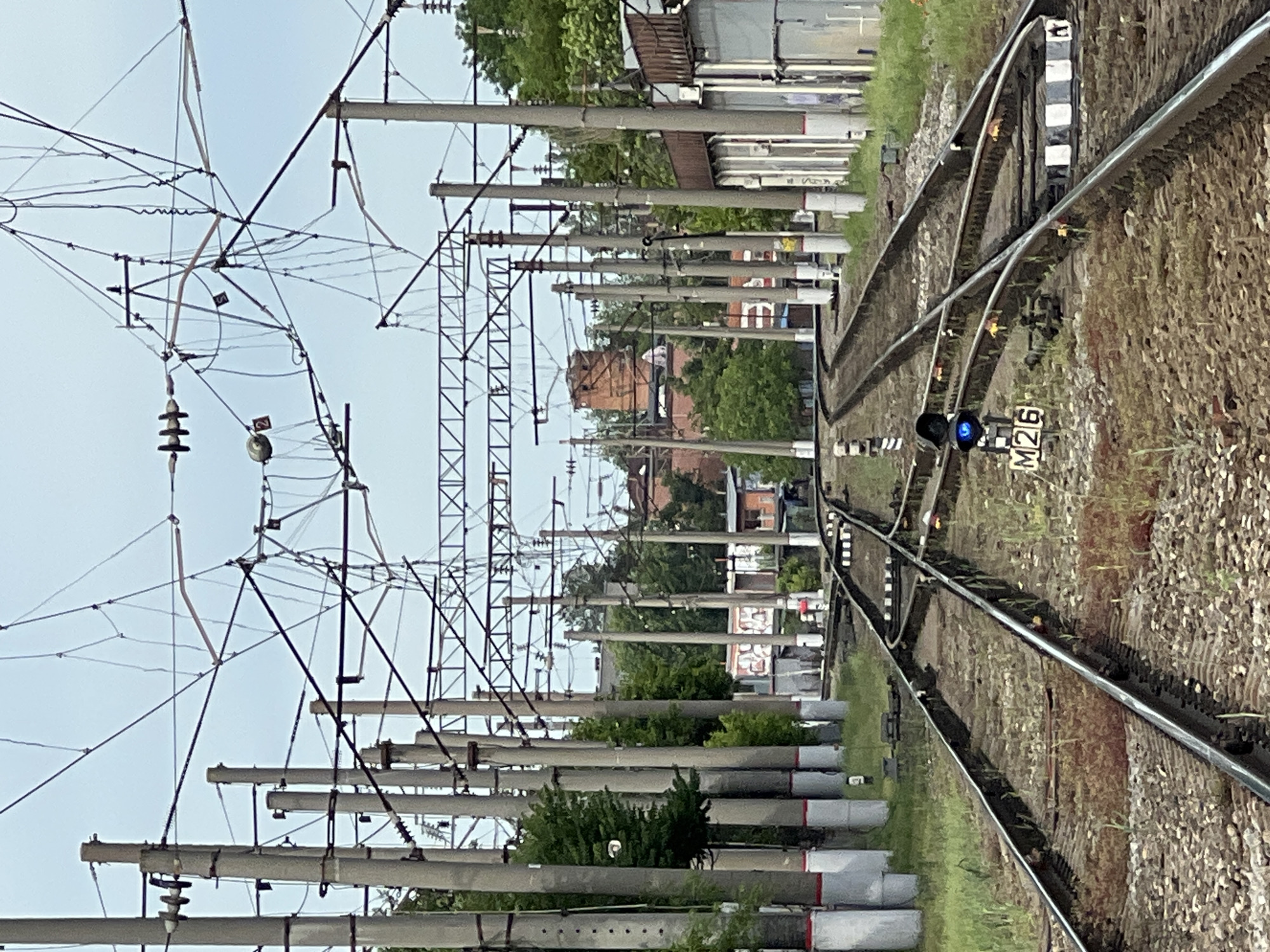 The image depicts a railway scene, oriented vertically. The tracks curve slightly to the right, with a junction visible in the middle ground. To the right of the main track, a blue signal light and a sign reading "M26" are observed. Overhead, a complex network of overhead lines and supporting poles dominates the view. A line of concrete utility poles runs parallel to the tracks. In the background, hints of urban structures are visible, including a brick building and a series of industrial buildings. This appears to be Krasnodar, Russia.