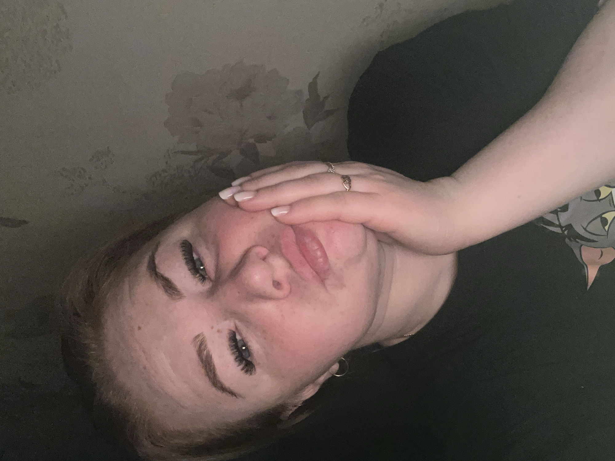 An adult female is depicted in a close-up, dimly lit indoor setting, presumably within a residence in the city of Chaikovsky, Russia. Her fair complexion exhibits flushed cheeks and scattered freckles. Her reddish-brown hair is pulled back, partially obscuring the top of her head. Her eyes, adorned with prominent dark lash extensions, are partially closed and directed towards the camera. Her lips are puckered in an expressive pose. A small gold hoop earring is visible on her left earlobe. Her right hand is raised, with manicured nails featuring a French tip design, resting against her right cheek. Two rings, one with a textured band and another with a small decorative knot, are present on her right ring finger. She wears a dark-colored top, possibly a t-shirt, with a partially visible graphic of a cartoon cat's face (resembling Tom from Tom and Jerry) on the lower right. A thin gold chain is visible around her neck. The background is a muted, light brown or grey wall adorned with a subtle, lighter-toned floral or botanical pattern, indicative of wallpaper.