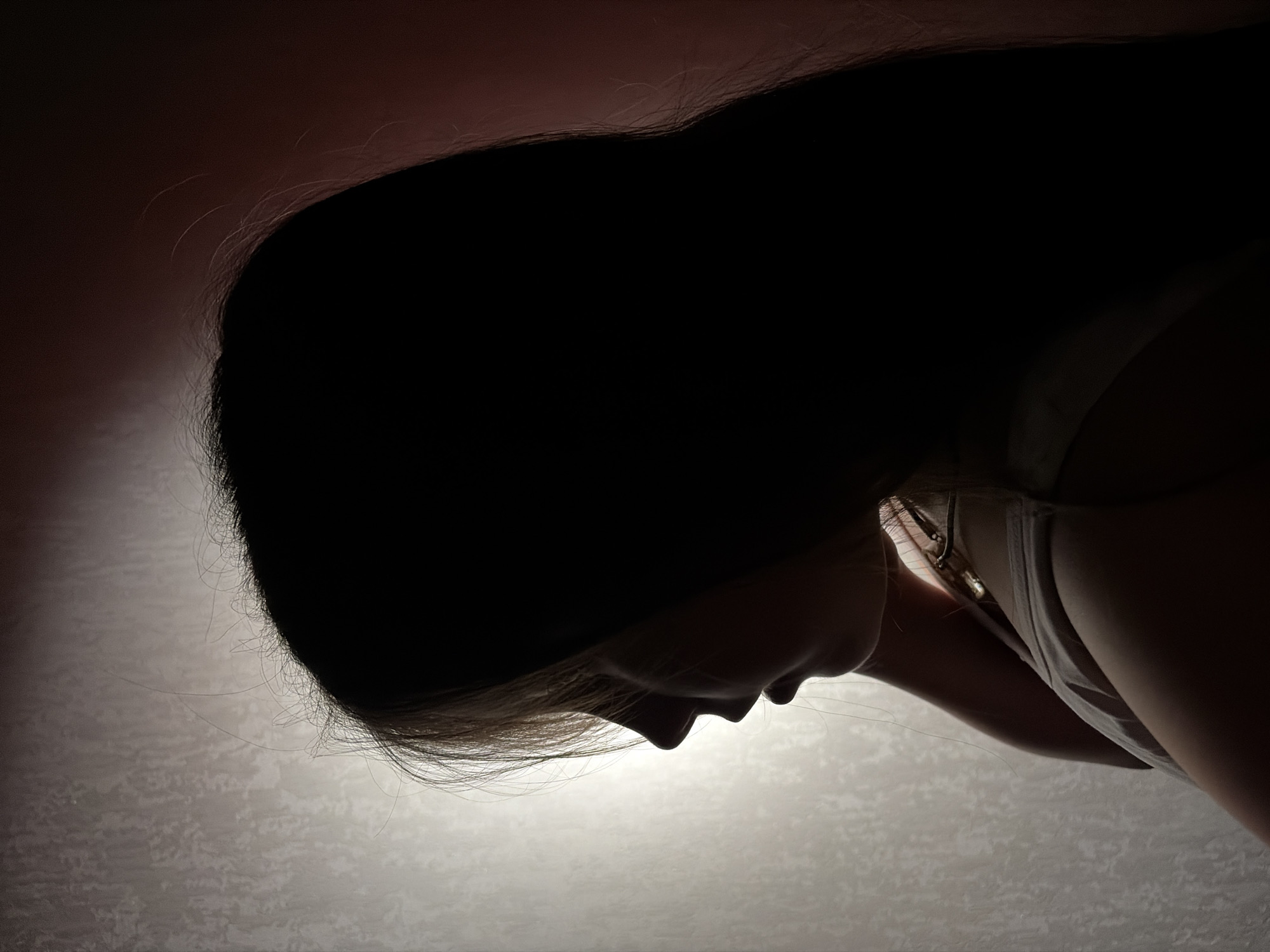 The image shows a person with long, dark hair, head bent down, facing downward. A white top is visible. The background is a gradient of dark to light, suggesting a dim light source from below. The location is Unknown, Unknown.
