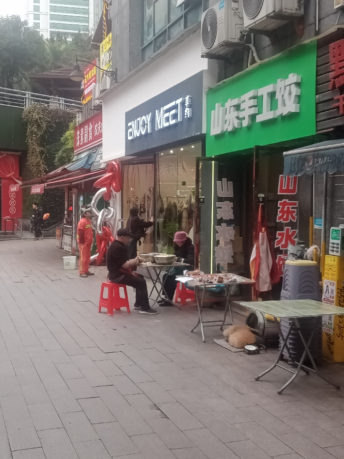 A street scene features several people and a dog. Two individuals are seated at tables preparing food, with red stools nearby. Two workers in orange uniforms stand further down the street. Silver balloons add a decorative element. A light brown dog is curled up asleep beside one of the tables. Shop signs in Chinese are visible above, including "ENJOY MEET" and a green sign with white lettering. Air conditioning units are mounted high on the buildings.