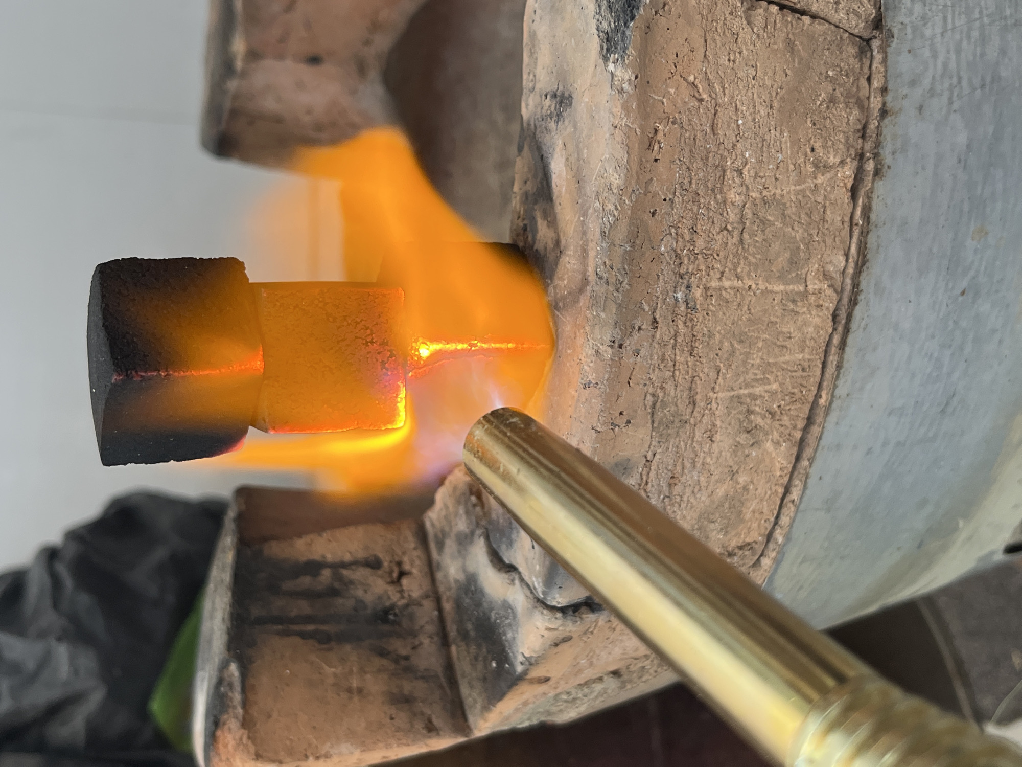 A close-up shot depicts a metal object being heated with a golden-colored torch inside what appears to be a kiln or furnace constructed of a rough, light brown, porous material. The metal object, which appears to be a bolt or similar hardware, is glowing orange-hot in the area being directly heated by the torch. The non-heated portion of the bolt is blackened. Flames from the torch engulf the metal piece. The setting seems to be a workshop environment. There's no indication from this image that this scene is located in Bac Nha Trang Ward, Vietnam.