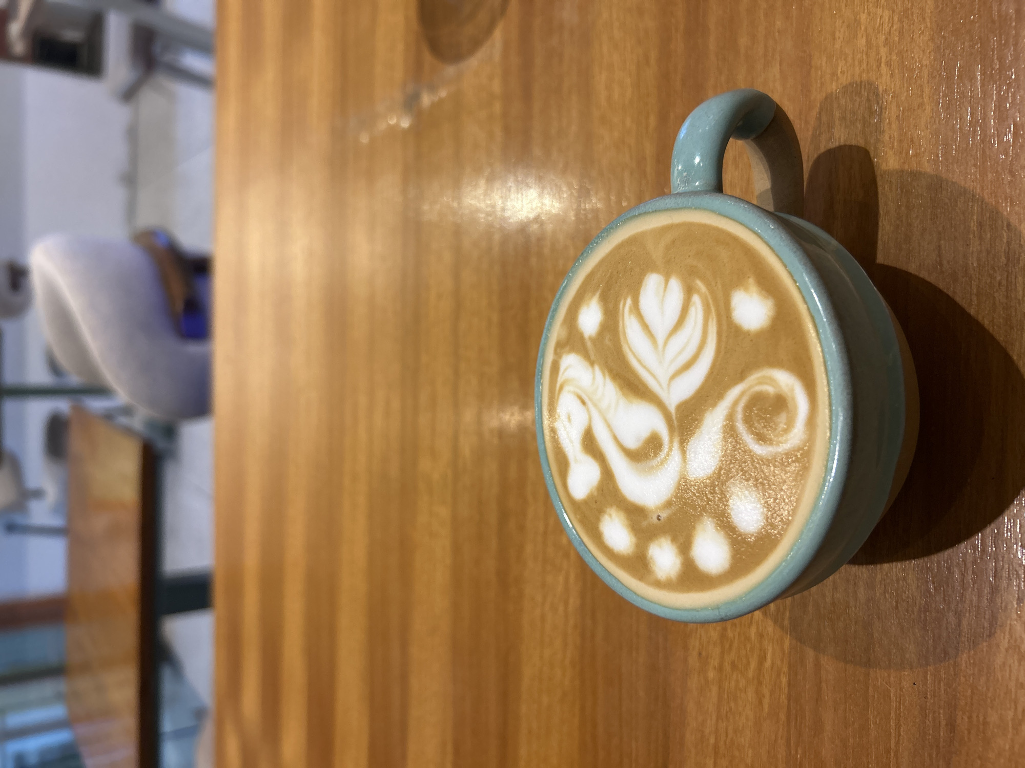 The primary subject of the image is a light teal-colored ceramic coffee mug, viewed from above, containing a beverage topped with intricate latte art. The art, created with white milk foam against the rich brown coffee, depicts a stylized design resembling a swan or mythical creature with flowing lines and a floral element, accompanied by several small, round white dots.
The mug rests on a smooth, polished wooden table, which occupies the majority of the foreground and midground. The table exhibits a warm, medium-brown hue with subtle vertical wood grain patterns and some reflective highlights on its surface. The mug casts a soft yet distinct shadow on the table to its right side.
In the blurry background, to the left of the table, an out-of-focus indoor environment is visible. This indistinct setting includes a soft, light-colored object, possibly a piece of furniture like a chair, and various dark and light shapes and lines that suggest a cafe or an interior room.
The lighting in the scene is soft, enhancing the textures of the latte art and creating depth through the shadows. No text is visible in the image. The scene captures a still life moment focused on the aesthetic appeal of a crafted beverage.