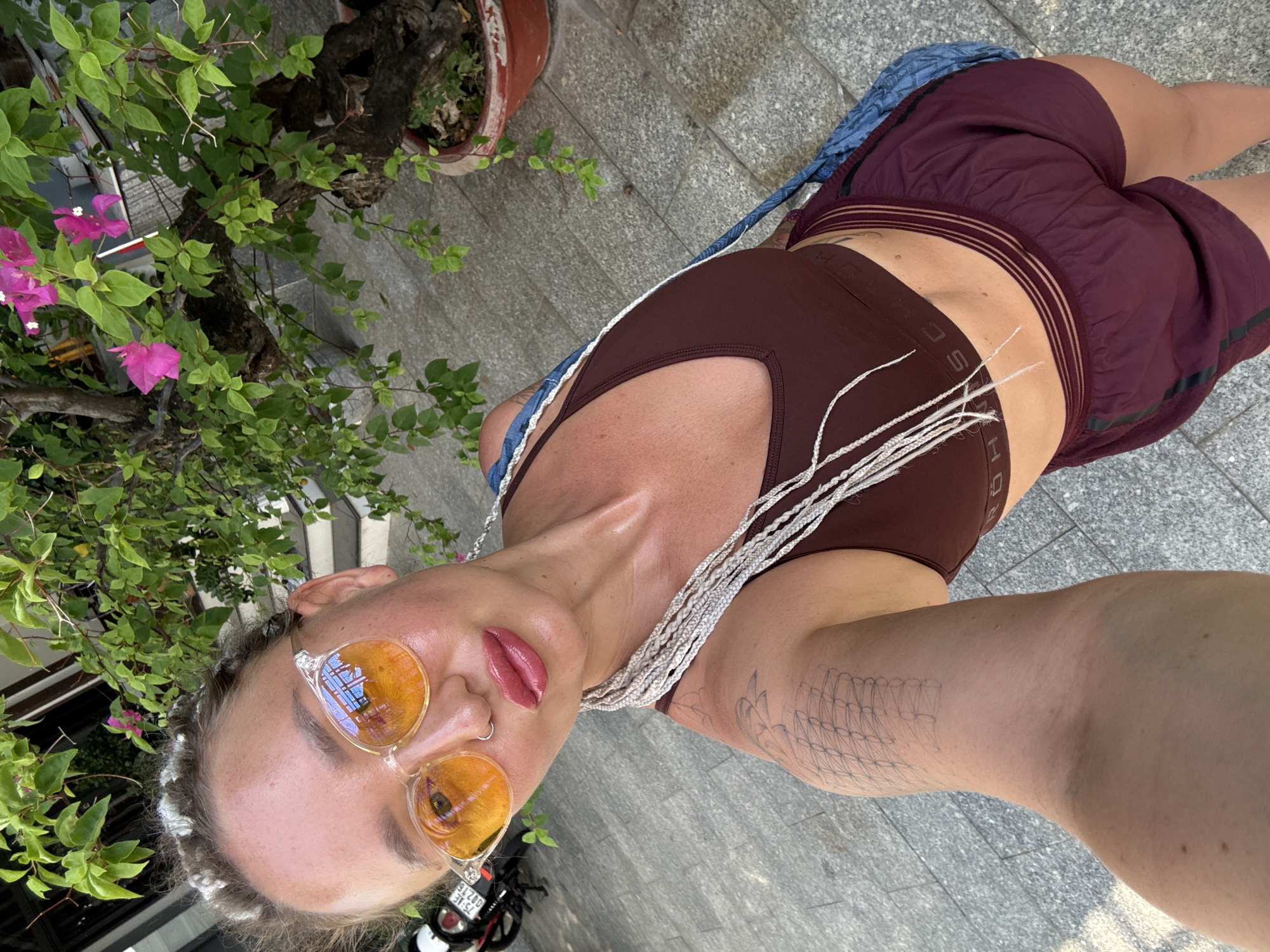 A woman with blonde braided hair and orange-tinted sunglasses is taking a selfie outdoors. She is wearing a brown sports bra and maroon shorts. Her left arm, visible in the foreground, has a geometric tattoo. Behind her, there is a tree with pink flowers and green leaves, and a paved walkway. A motorcycle is partially visible on the left side of the image. The scene appears to be in a city, possibly in the Bac Nha Trang Ward of Vietnam, suggested by the architecture and vegetation.