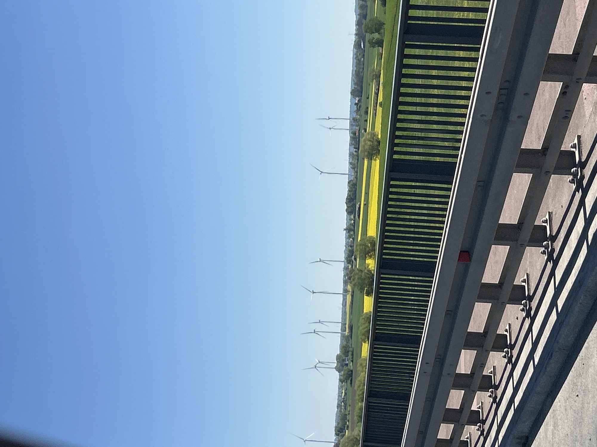The image shows a view from a bridge in Kończewice, Poland. A blue sky fills the left side of the frame. On the right is a section of the bridge's guardrail, constructed from gray steel and concrete. Beyond the guardrail, a landscape stretches into the distance. A field is striped with green and yellow, indicating different crops or vegetation. Several wind turbines are visible on the horizon, their blades adding a sense of scale to the landscape. The scene is well-lit, suggesting a sunny day.