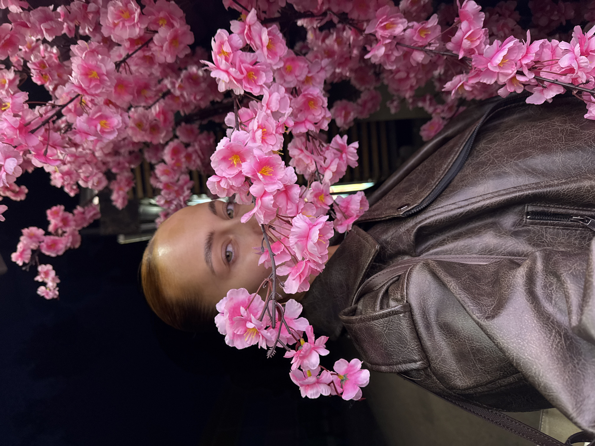 The image features a close-up portrait of a person partially obscured by a dense arrangement of artificial pink cherry blossoms, likely taken in Izhevsk, Russia. The central subject is a woman, whose right eye, forehead, and part of her nose are visible as she peers through the flowers, looking directly at the viewer. Her light brown hair is visible on the side of her head. She is wearing a dark brown, textured jacket, which appears to be made of faux leather, with visible zippers and pocket details on the shoulder and chest area.
The vibrant pink blossoms dominate the foreground and background, creating a soft, ethereal frame around the woman. The flowers are uniformly colored with yellow centers, suggesting they are artificial decorations. They are arranged on dark branches, filling the top and right side of the frame, with some individual blossoms falling closer to the viewer. The background behind the flowers is predominantly dark, almost black, indicating either a nighttime setting outdoors or a dimly lit indoor space. The lighting highlights the woman's face and the details of the flowers and jacket. The overall mood is intimate and aesthetic. No text is visible in the image.