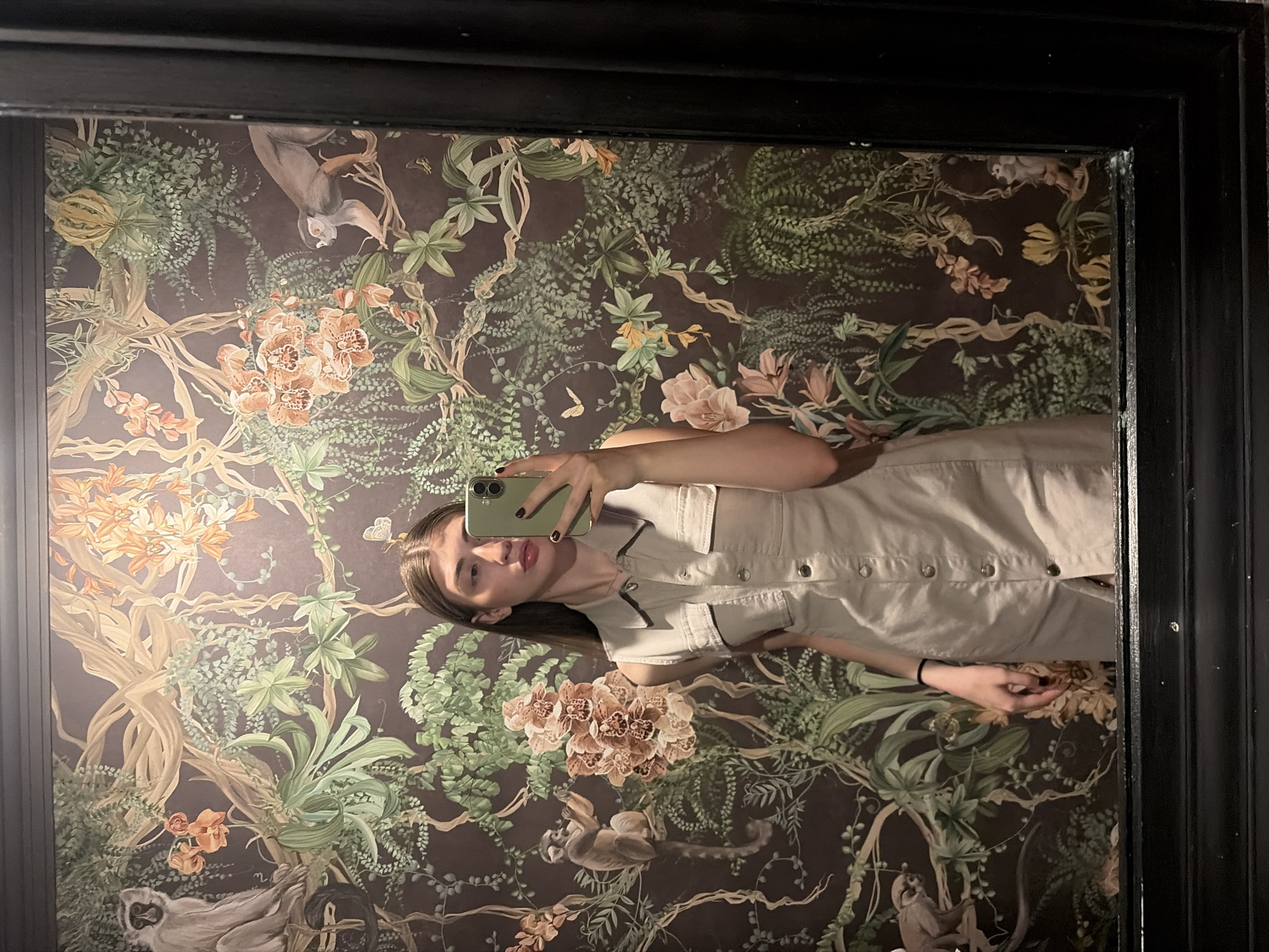 A young woman is captured in a mirror selfie, positioned centrally within a dark, ornate framed mirror. She has long, straight brown hair, parted in the middle, and is looking towards her smartphone. She is wearing a light beige or khaki-colored sleeveless dress with a collar, two chest pockets with buttoned flaps, and a row of dark buttons running down the front. Her right hand holds a light olive-green smartphone, obscuring part of her face, with dark nail polish visible on her fingertips. A thin black hair tie is on her left wrist.
The background, reflected in the mirror, is a richly detailed, dark brown wallpaper featuring a lush tropical jungle motif. Prominent elements include intertwining green vines, large green leaves, and ferns. Scattered throughout are clusters of orange and peach-colored orchids, along with other lighter-hued tropical flowers and smaller butterflies. Several monkeys are depicted within the foliage; one larger monkey is seated on a branch in the upper-left, another smaller monkey appears to be holding a plant stem near the center-left, and a third monkey is visible climbing on the right side of the wallpaper.
The mirror itself has a thick, dark, possibly black, wooden frame with some decorative molding. A slightly lighter grey/brown molding is visible above the top of the mirror frame, and a dark wooden base or lower frame is present at the bottom edge.
If located in Krasnoyarsk, Russia, this highly decorative interior suggests a commercial establishment such as a restaurant, bar, or a refined public restroom within a hotel or cultural venue, characterized by its exotic botanical wallpaper design.