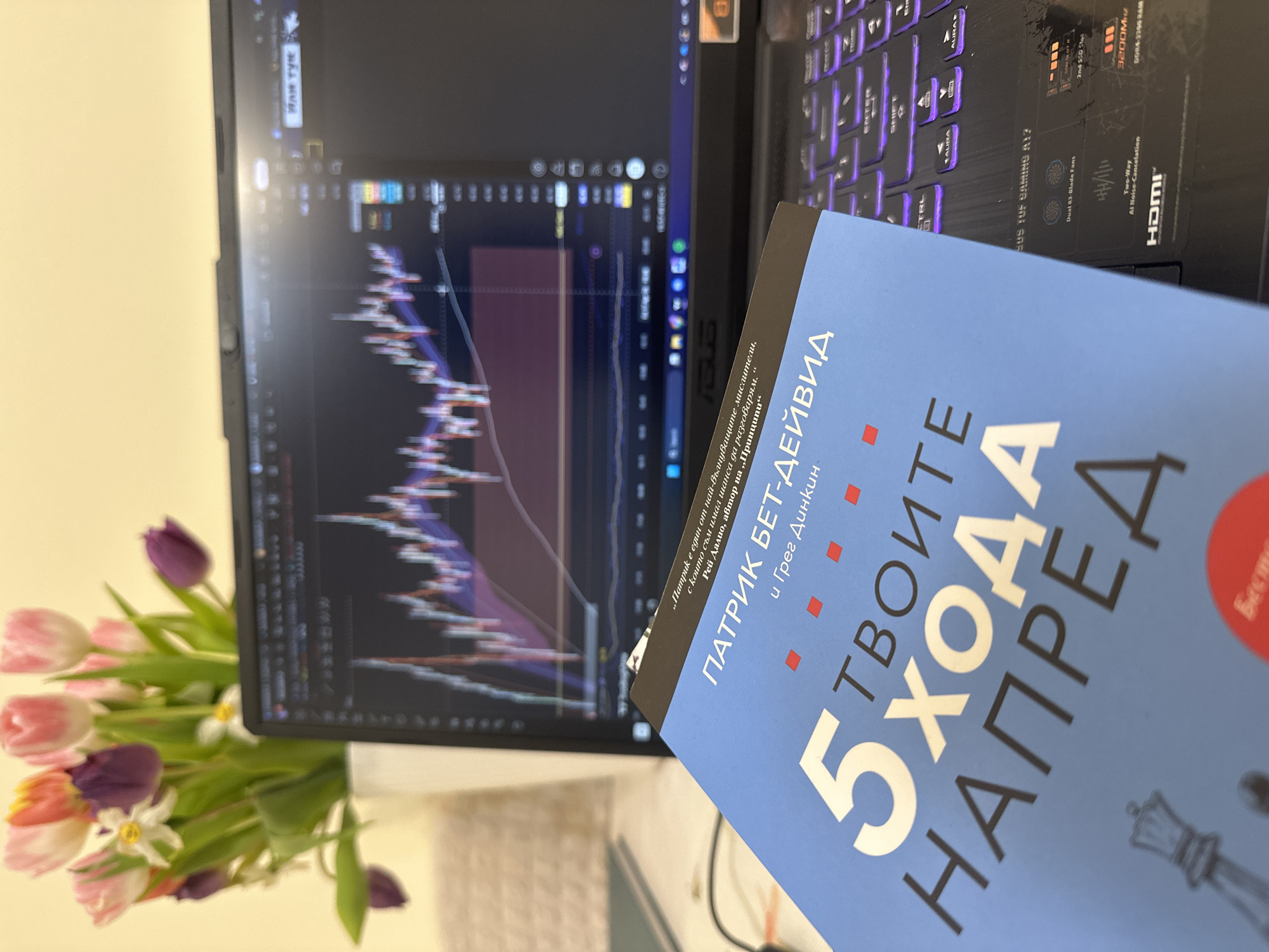 Here's a detailed description of the image:
**Subjects and Objects:**
* A laptop with a chart displayed on the screen (likely a financial chart indicating stock prices or trading data).
* A book titled "5 Steps Ahead" (translated from Bulgarian).
* A vase of colorful tulips.
* A laptop keyboard with purple backlighting.
**Location:** The scene appears to be indoors, possibly in a home or office setting. There's no immediate indication of Varna, Bulgaria from the image itself.
**Environment and Setting:**
* The background is light and fairly nondescript.
* The lighting appears to be ambient or artificial.
**Details:**
* The book is resting on the laptop keyboard.
* The flowers are positioned next to the laptop screen.
* The laptop is an ASUS TUF Gaming A17.