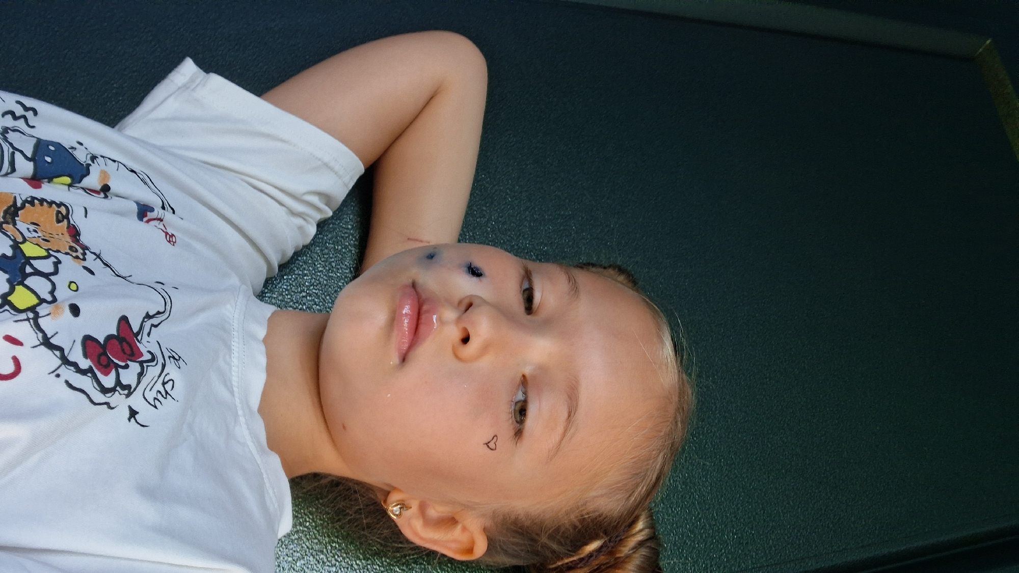 The media file is a portrait of a young girl lying down, her head tilted and resting on her arm. She is wearing a white t-shirt with a cartoon print featuring characters like Hello Kitty and what appears to be Donald Duck. The girl has blonde hair styled in braids and is looking towards the camera with a soft expression. She has some blue markings or smudges on her cheek, possibly from face paint or a temporary tattoo. A small black heart tattoo is visible on her left cheekbone. She is wearing a small hoop earring in her left ear.
The background is a textured, dark green surface, suggesting an indoor or shaded outdoor setting. The lighting appears to be natural, with soft illumination that highlights the girl's features. The overall mood is relaxed and intimate, possibly a candid moment captured during playtime or a casual outing. There is no visible text in the image itself, but the t-shirt has printed designs. The provided location context of Semey, Kazakhstan, offers a geographical setting for the image.