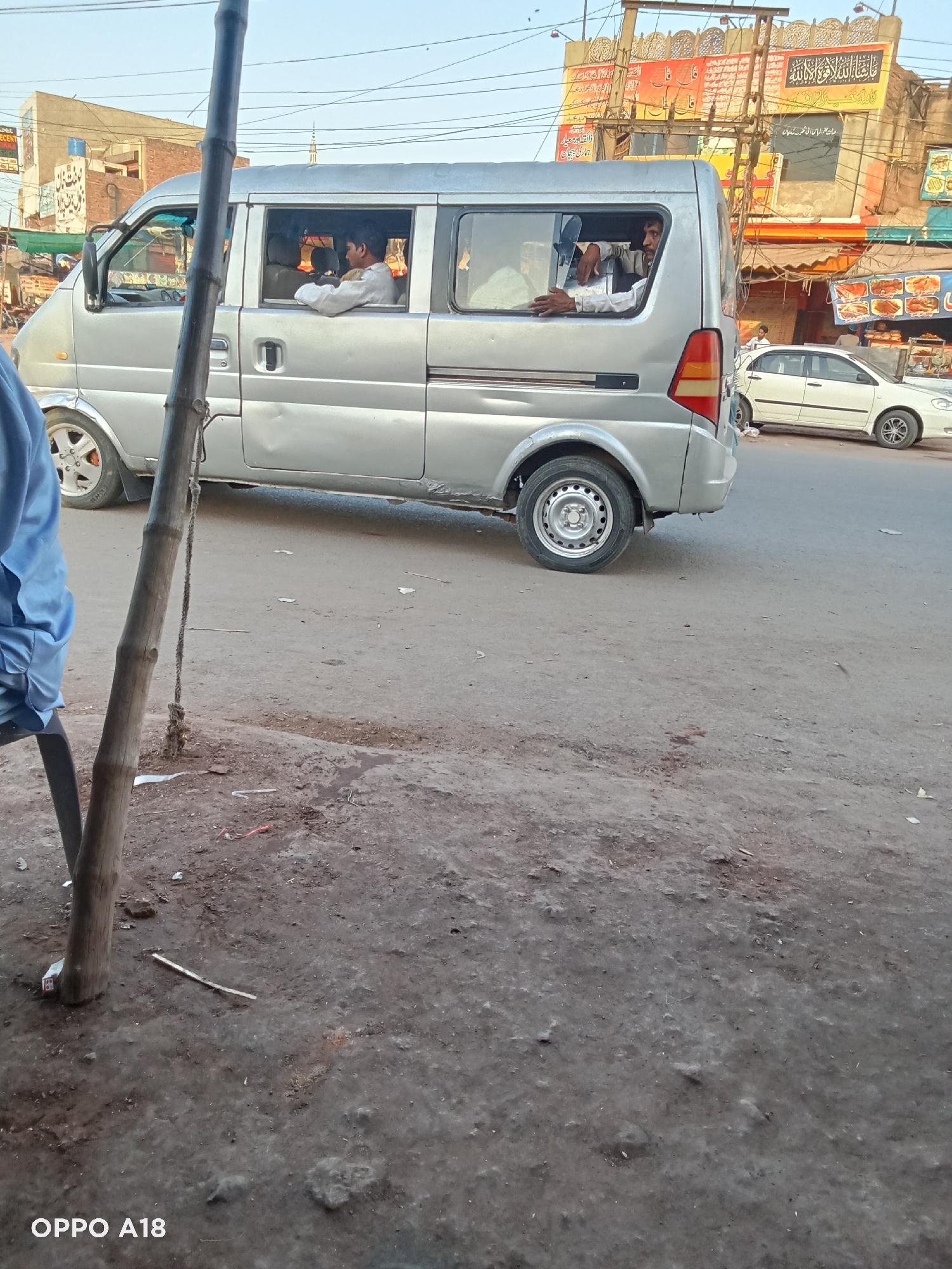 The image shows a silver van with three visible passengers on a street in an urban setting. The van is parked on the side of the road, and there is a white car parked further down the street. The background includes buildings with signs in what appears to be Arabic script and images of food. A wooden pole with a rope attached is in the foreground on the left side of the frame. The street surface is uneven and appears to be made of asphalt. The text "OPPO A18" is visible in the bottom left corner of the image.