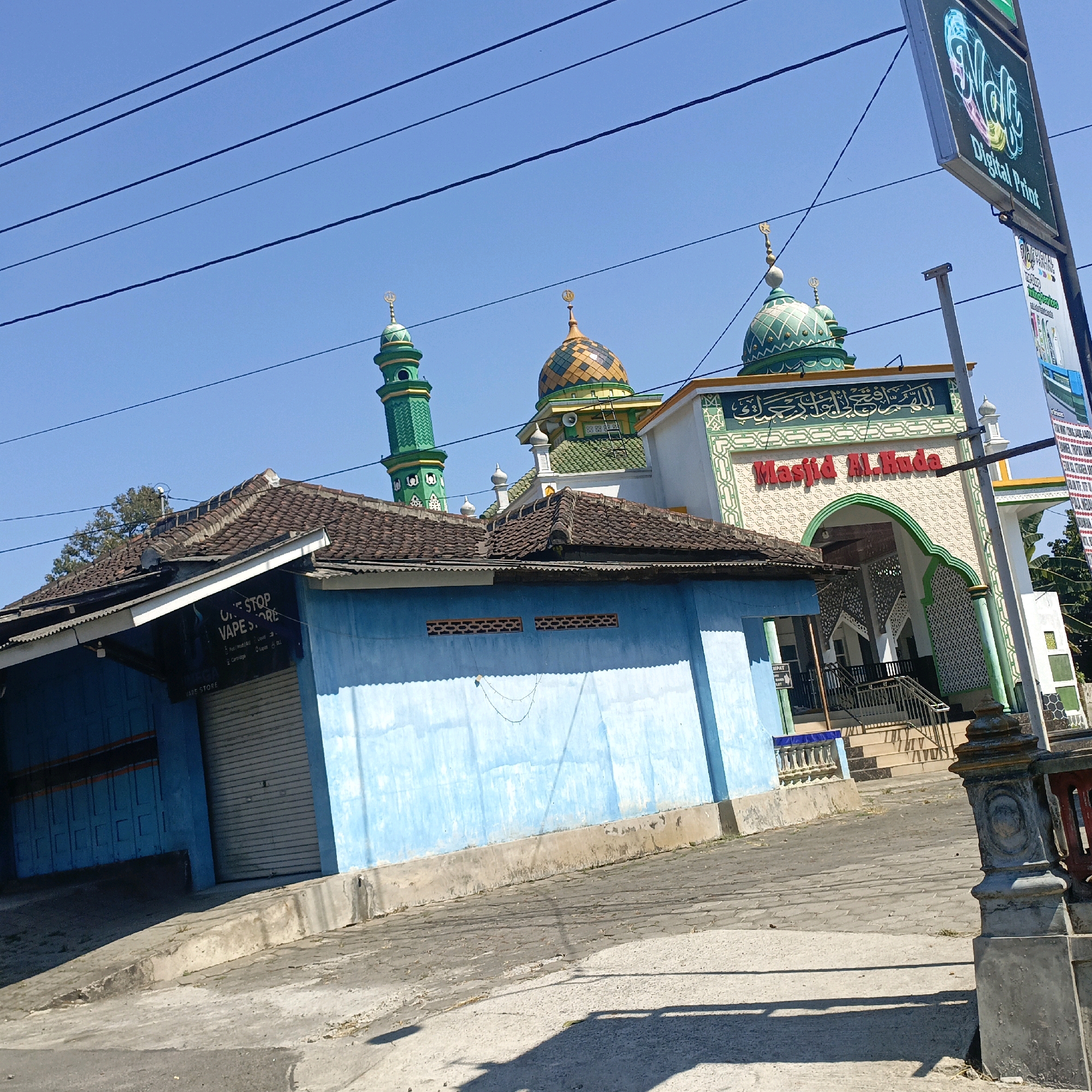 In the city of Kalibeji, Indonesia, a photograph captures a street view featuring a mosque and a vape store. The mosque, identified by Arabic inscriptions and the name "Masjid Al-Hada" in red lettering, is a prominent structure with green domes and minarets. Adjacent to the mosque is a light blue building housing "ONE STOP VAPE STORE," indicated by signage above a closed metal rolling door. The foreground shows a paved street and sidewalk, with electrical wires stretching across the clear blue sky. The scene appears to be a daytime shot, under bright sunlight, with shadows cast on the ground.